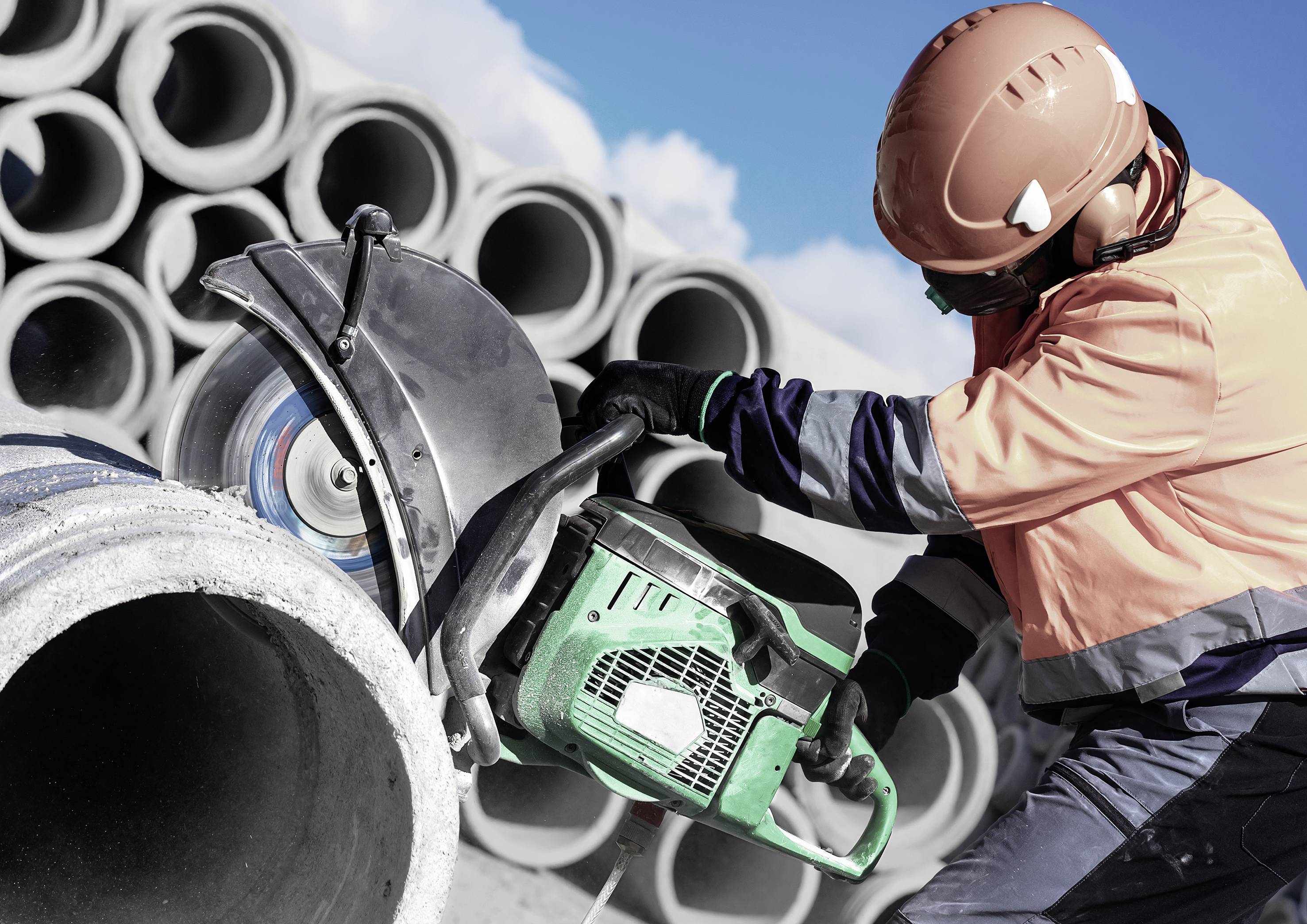 A worker is cutting a concrete pipe with a chainsaw, with stacked pipes in the background. The worker is wearing protective clothing.