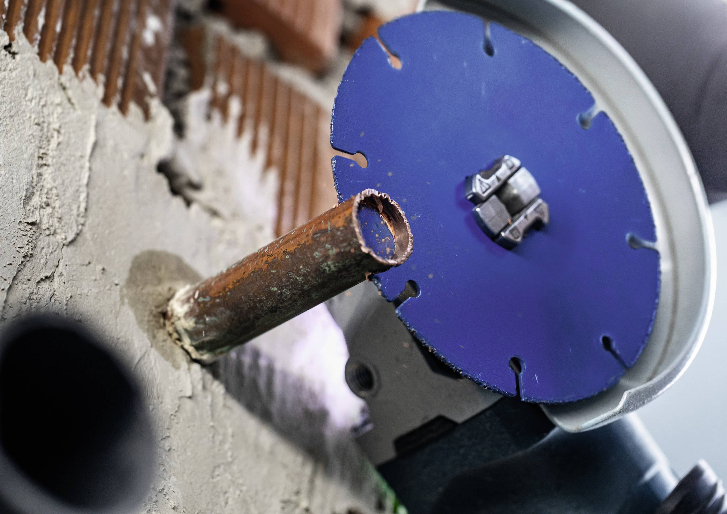 An angle grinder disc cuts through a metal pipe protruding from a concrete wall. Dust particles in the air.