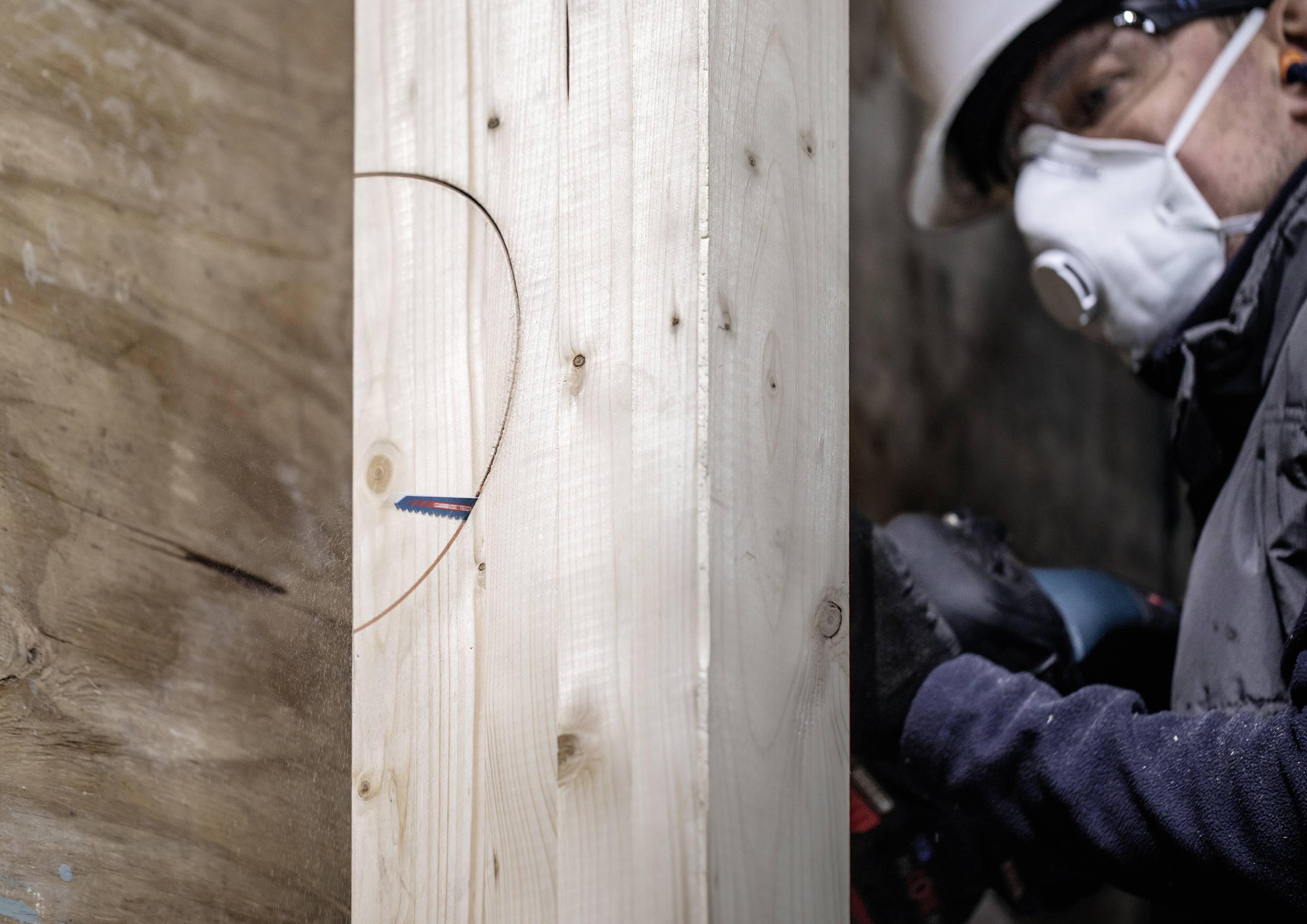 Worker wearing a protective mask drilling a hole in a wooden beam to prepare for construction work. Background shows construction site environment.