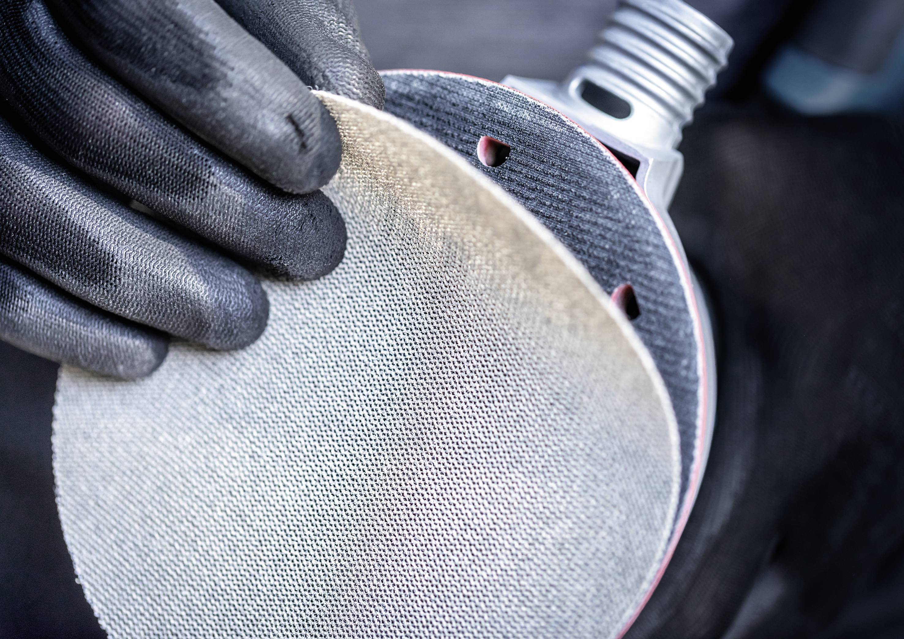A person wearing black gloves holds a round sanding pad in front of a grinding machine.