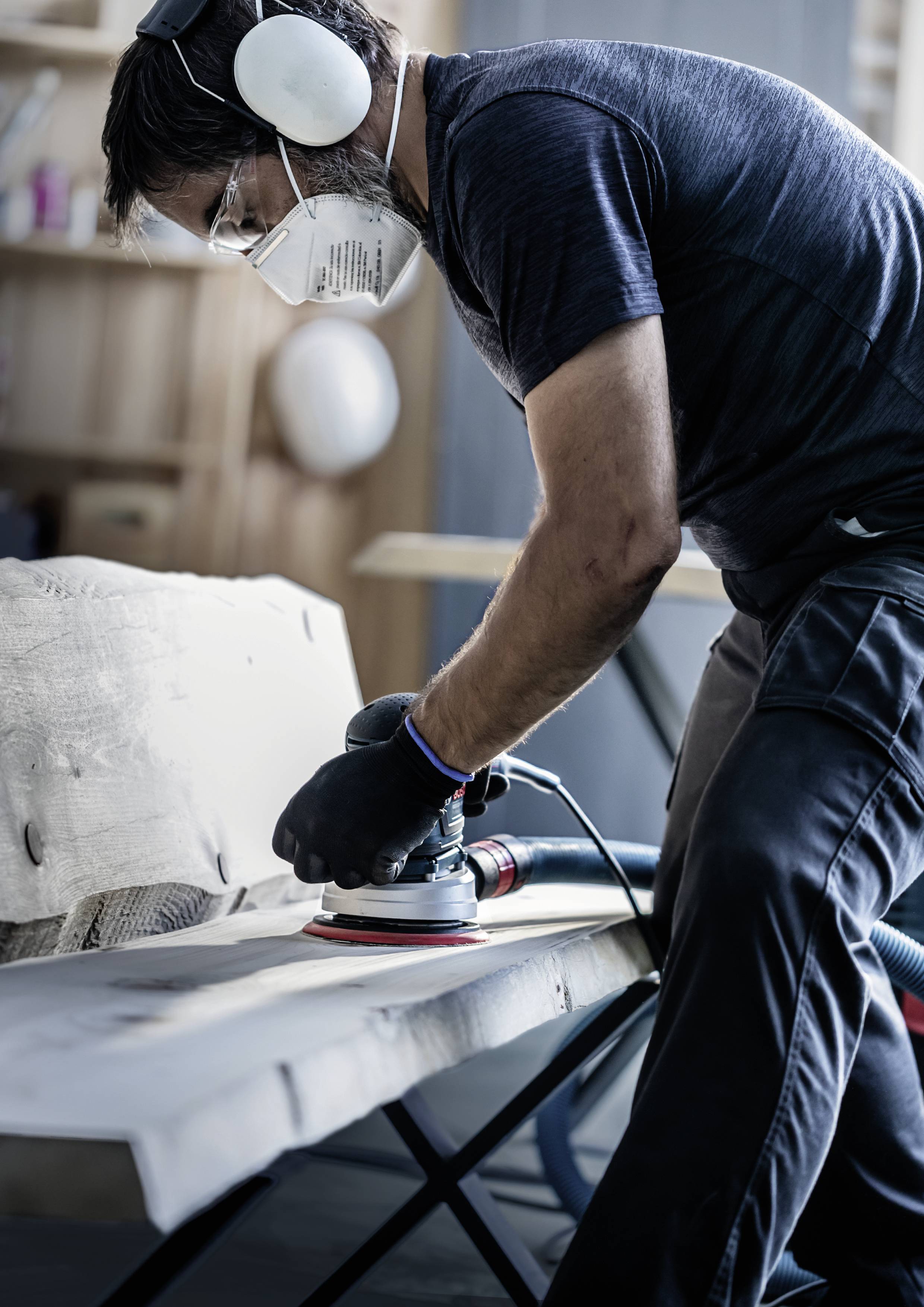 A person is sanding wood on a workbench, wearing ear protection and a respiratory mask in a workshop environment.