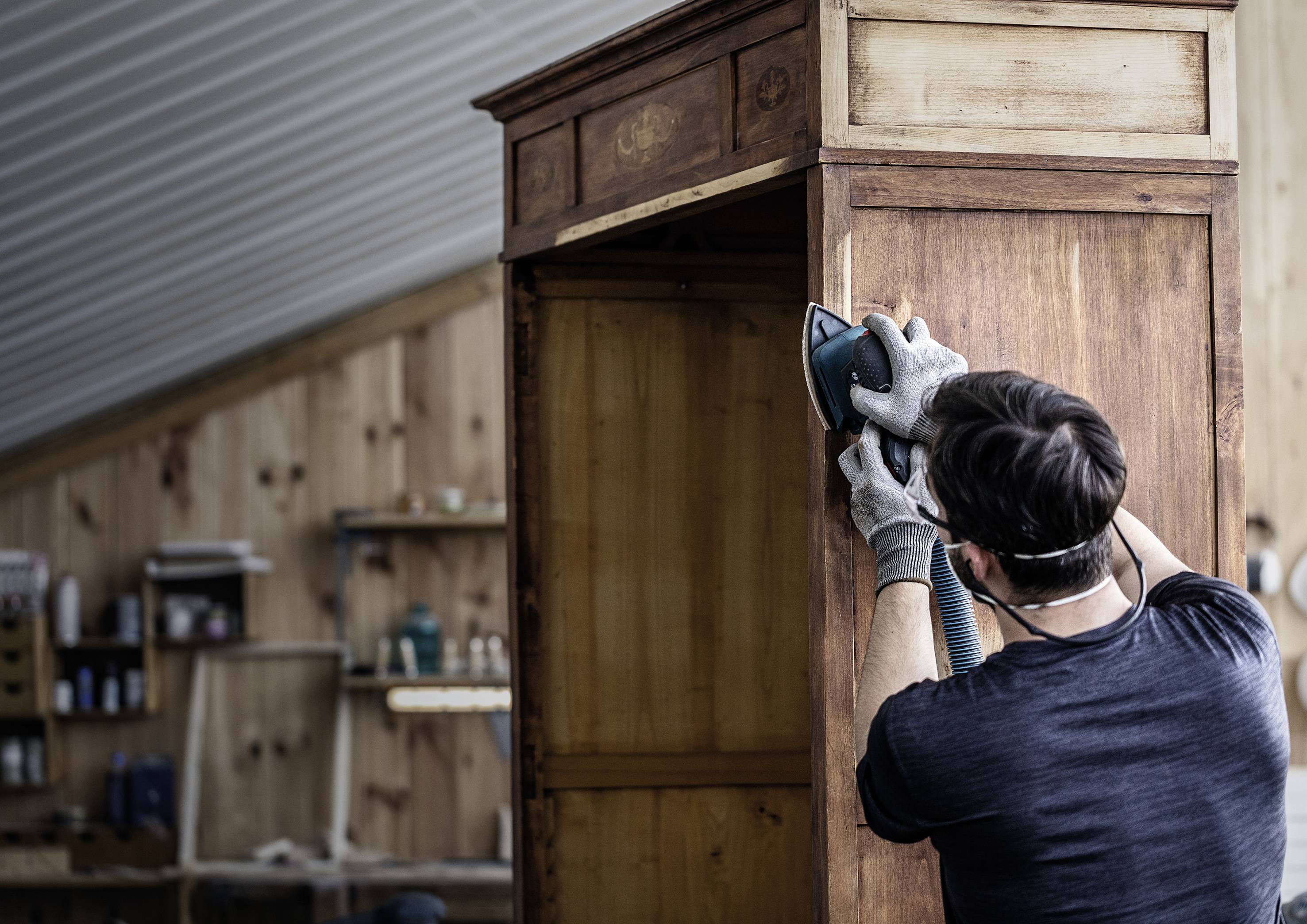 A person is sanding a wooden piece of furniture in a workshop. Shelves with tools and materials can be seen in the background.