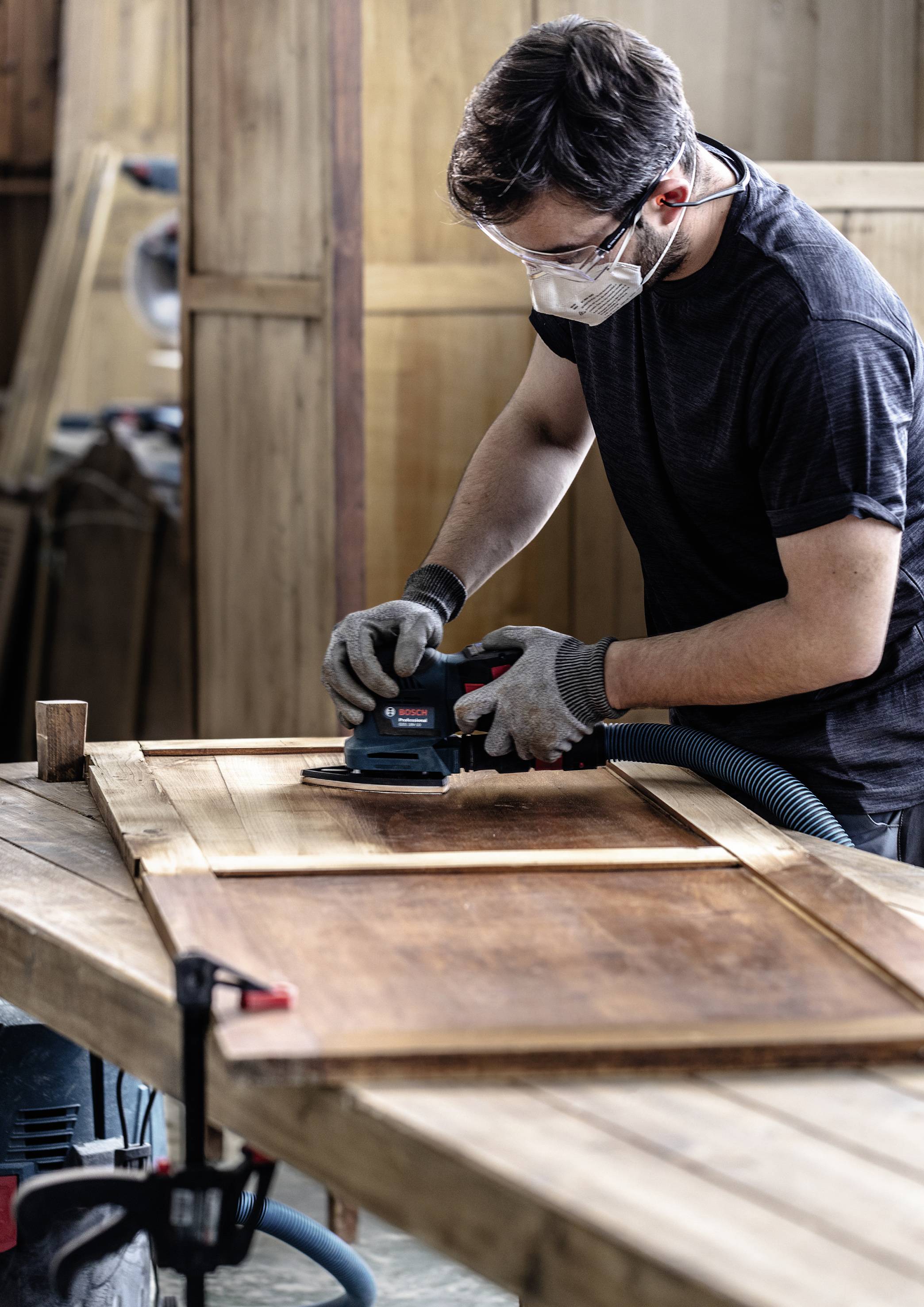 A craftsman wearing a respiratory mask is sanding a wooden door on a workbench in a workshop.