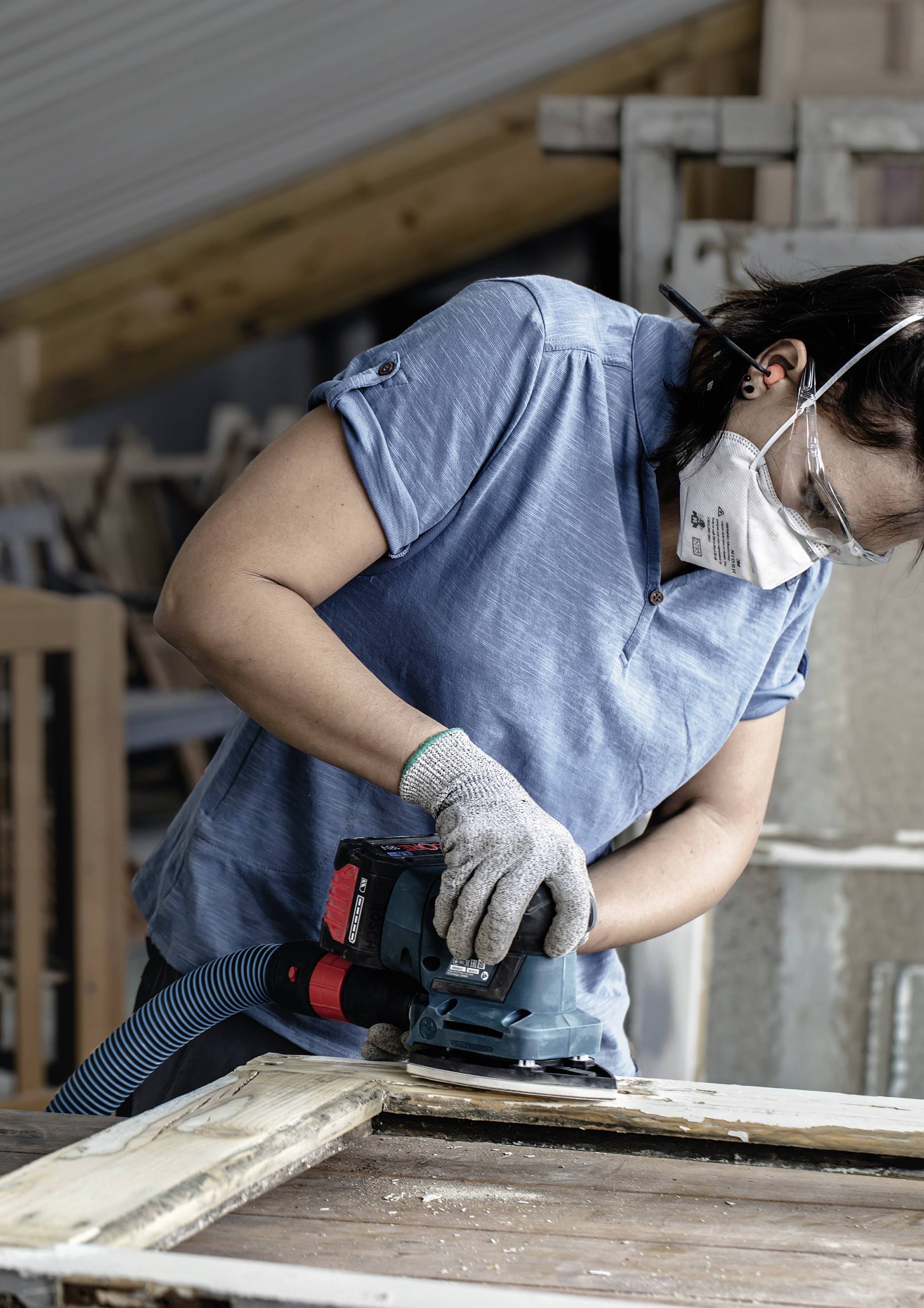 A person in a workshop is wearing a protective mask and sanding a wooden door with an electric sander. Wood shavings are falling off. Wooden furniture can be seen in the background.