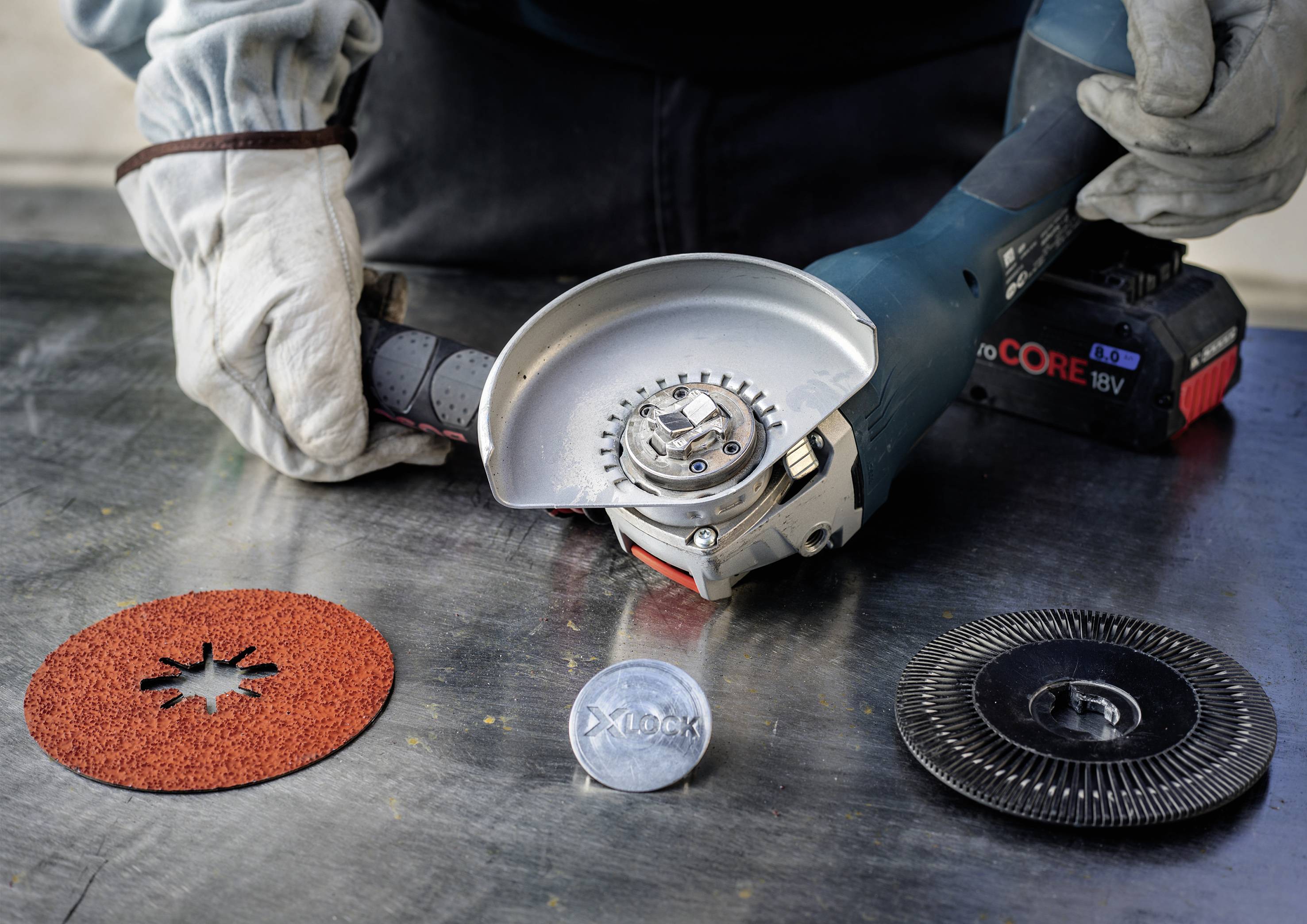 A person is wearing work gloves and holding an angle grinder on a table. Various grinding discs are laid out nearby for selection.