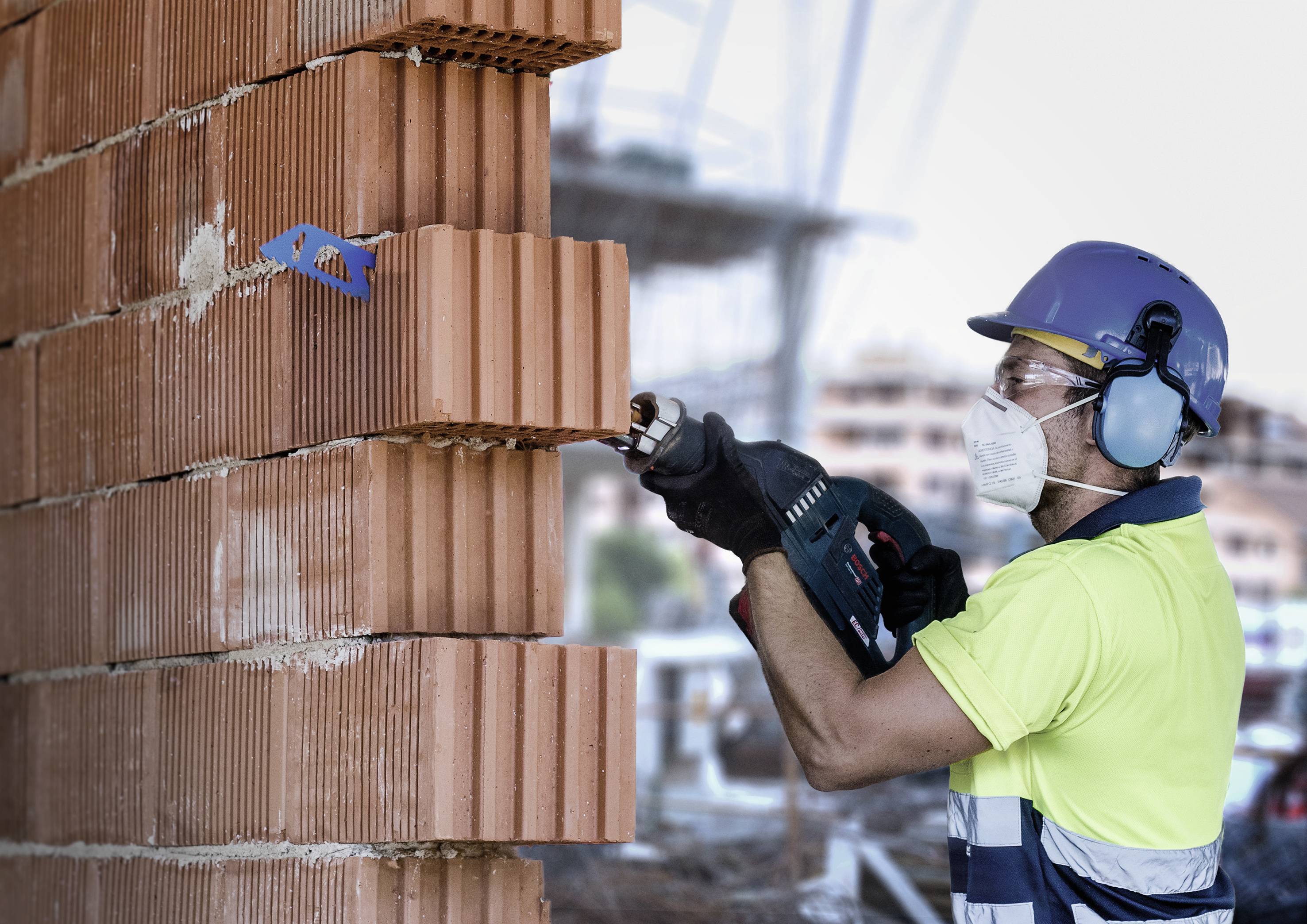 A construction worker wearing a hard hat and mask is drilling holes in a brick wall on a building site. Background blurred.