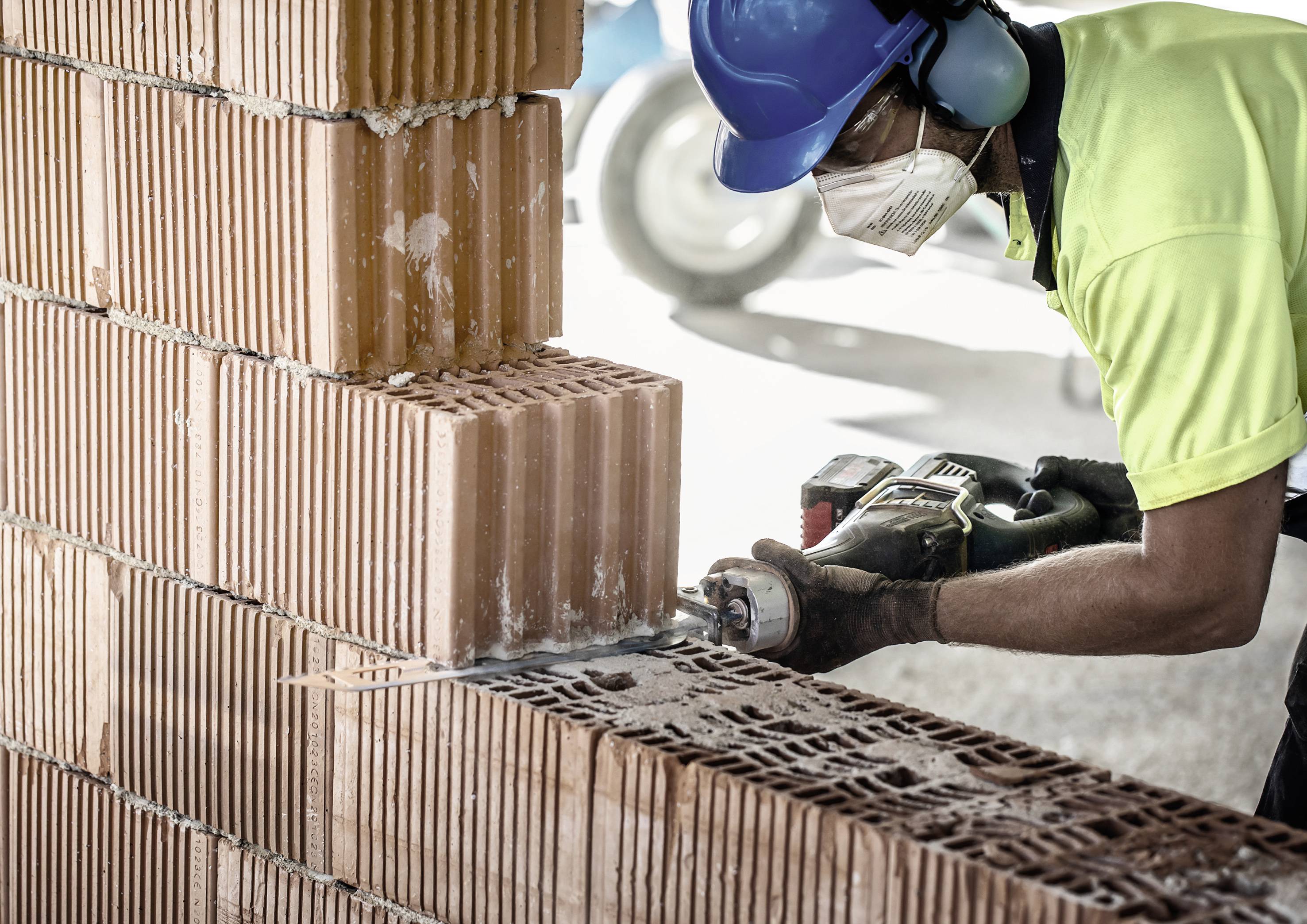 A construction worker cuts a red brick wall with an electric power tool. He is wearing a blue hard hat, ear defenders and a protective mask.
