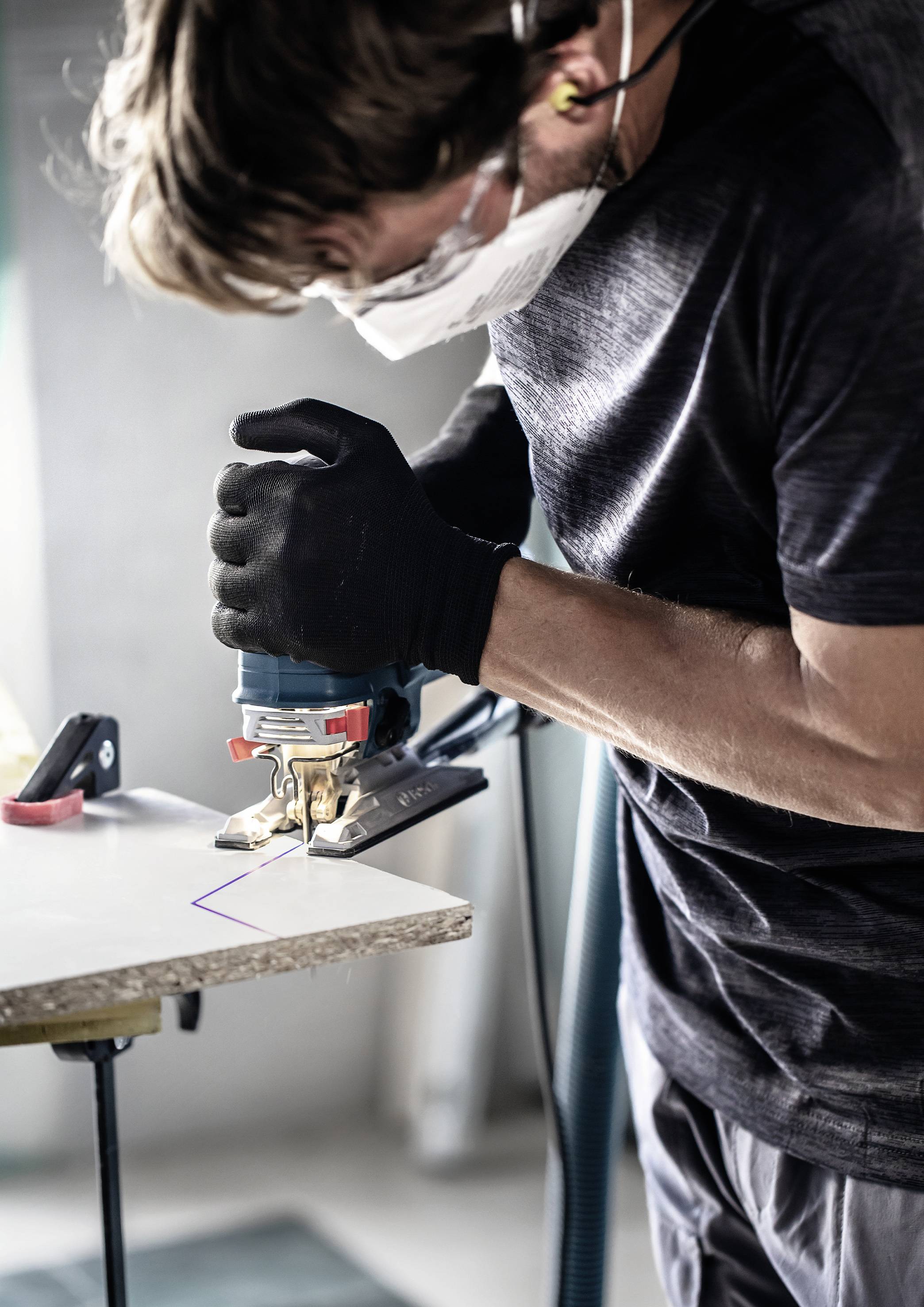 A person is wearing a protective mask and gloves and using an electric jigsaw to cut wood.