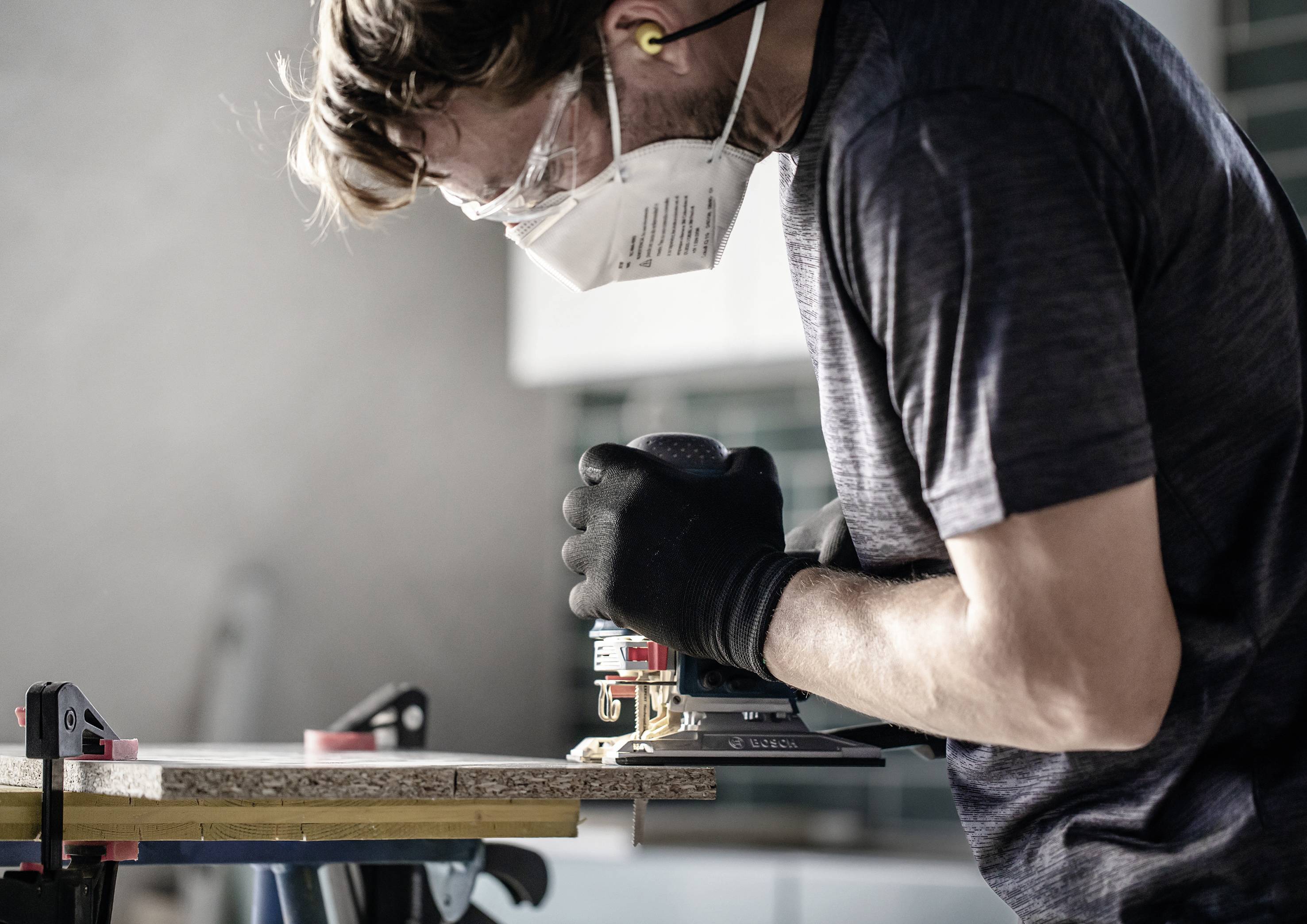 A man wearing a mask and ear protection is grinding a piece of wood with an electric grinding machine on a workbench in a workshop.