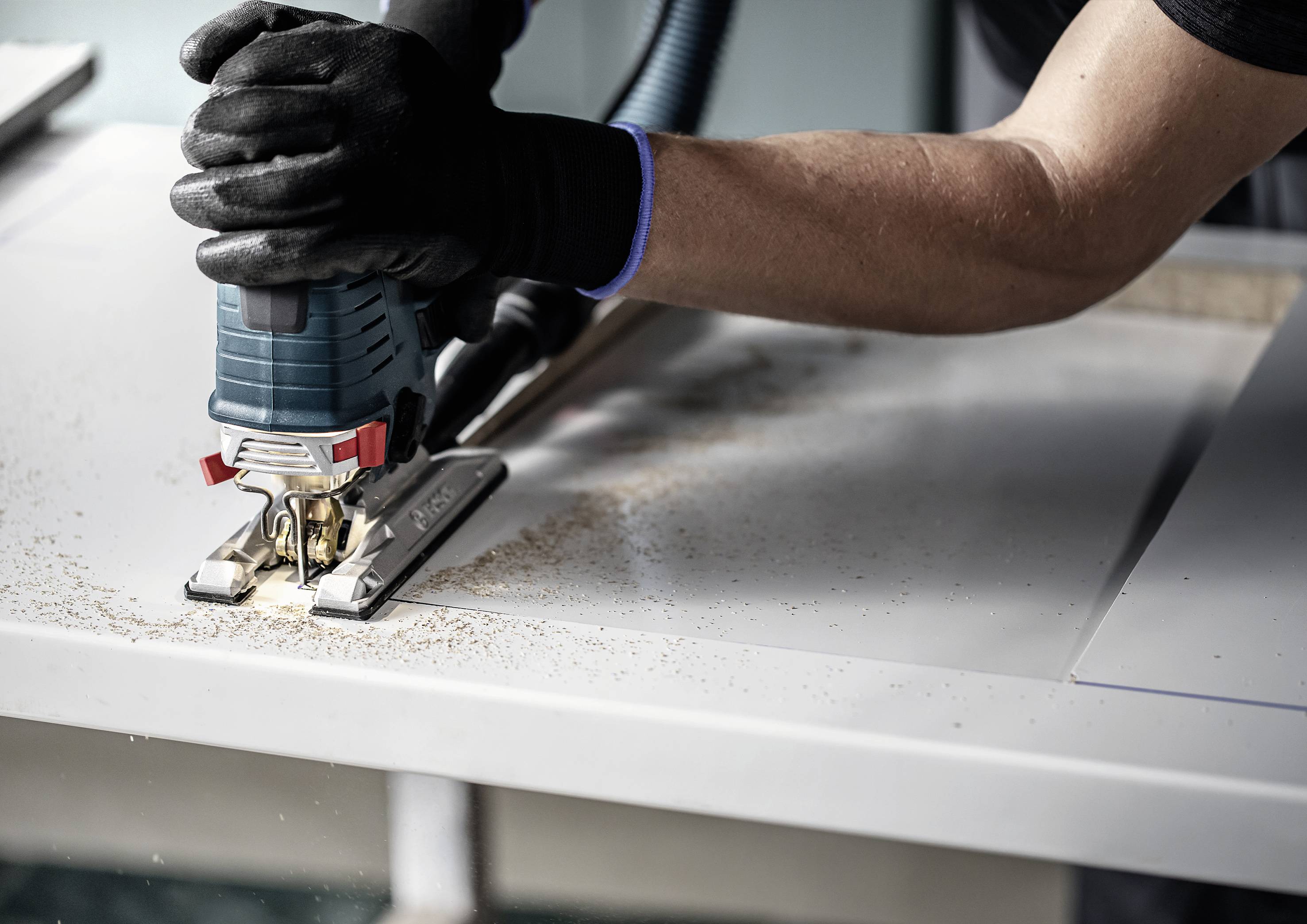 A person is working on a wooden board with an electric jigsaw. Sawdust is visible, and the person is wearing black gloves.
