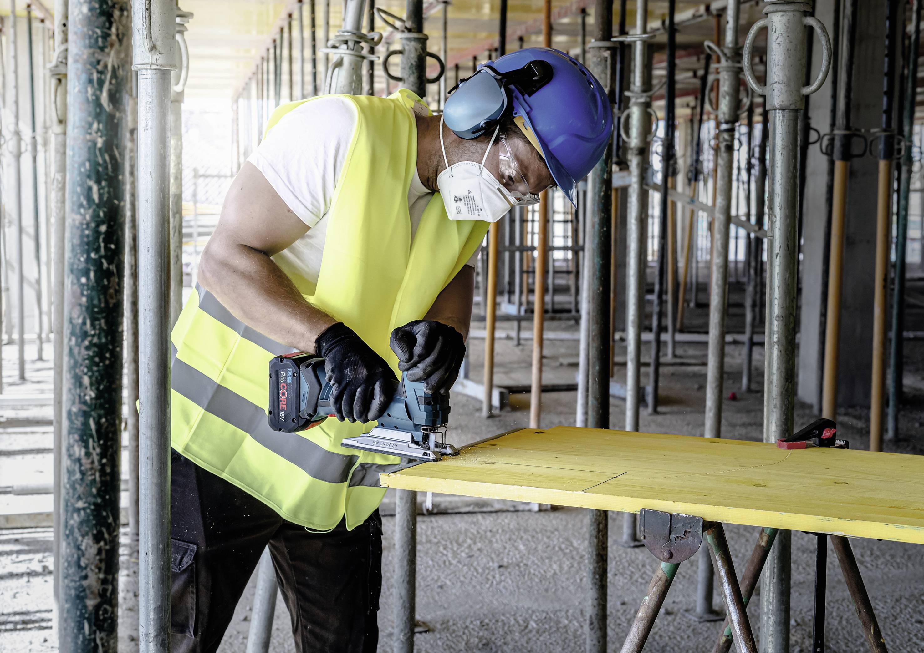 A worker wearing a hard hat and high-visibility vest is cutting a wooden board with a jigsaw on a construction site. Safety equipment is being worn.