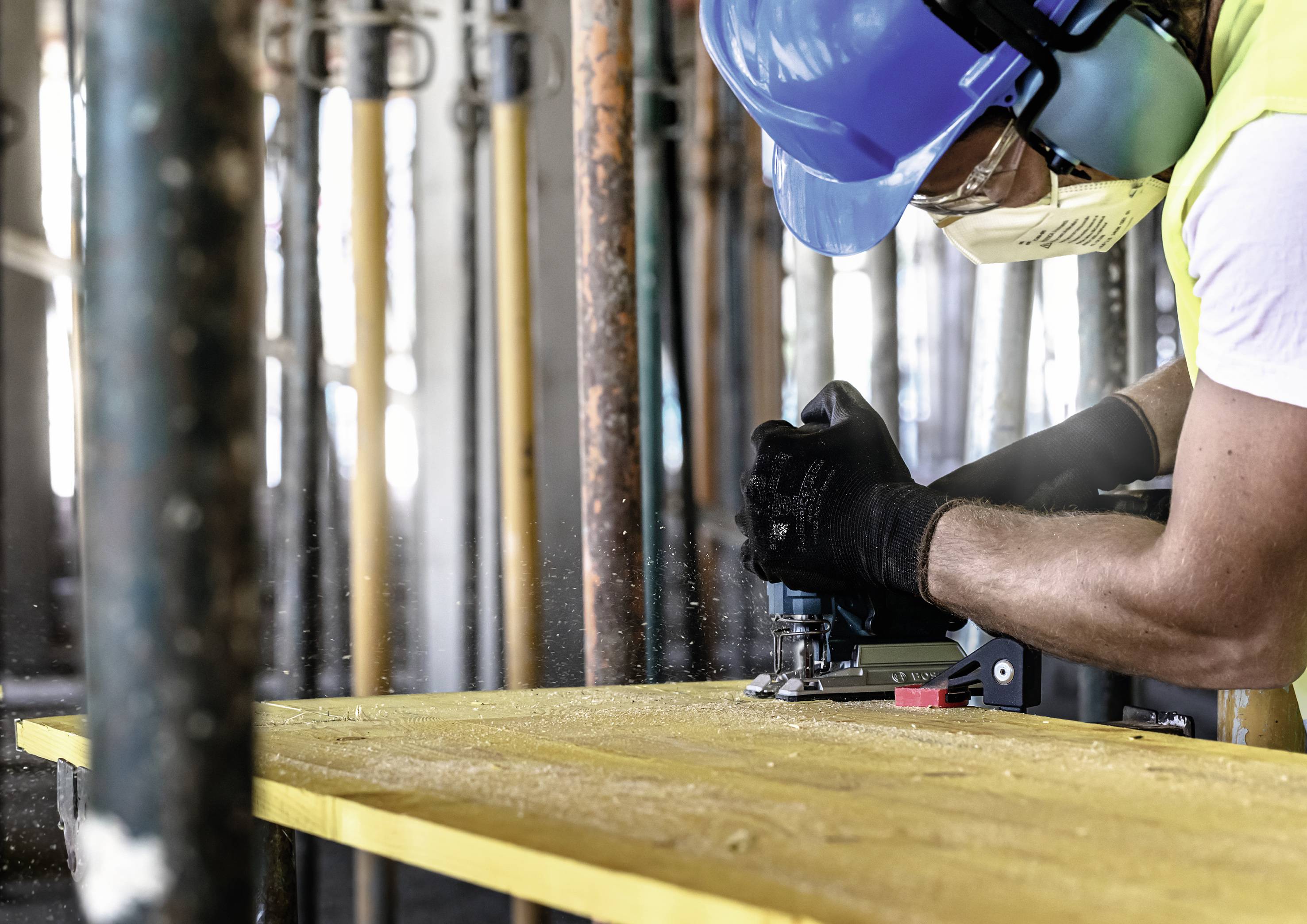 A person wearing a hard hat and safety glasses is using an electric saw to cut a yellow board at a construction site.