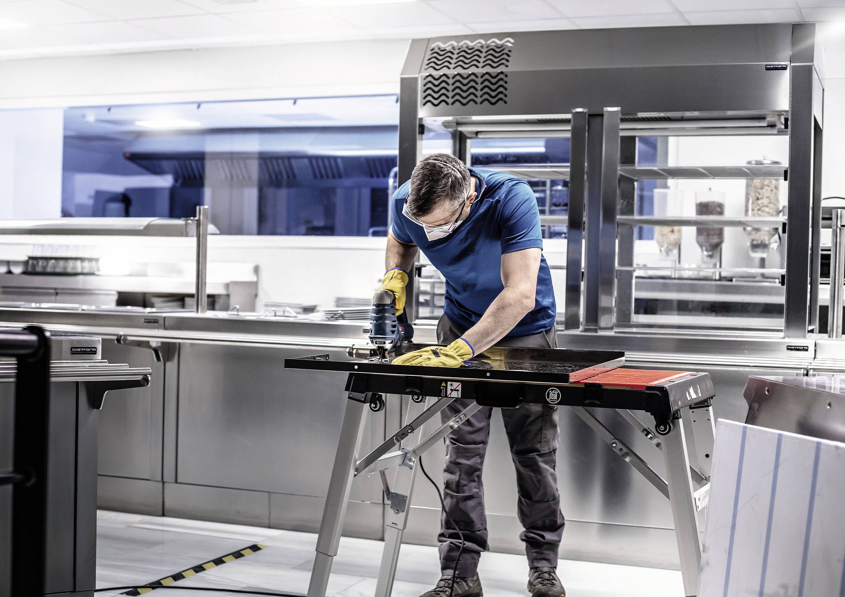A man is working with safety glasses and gloves on a workpiece in a modern, professional kitchen environment.