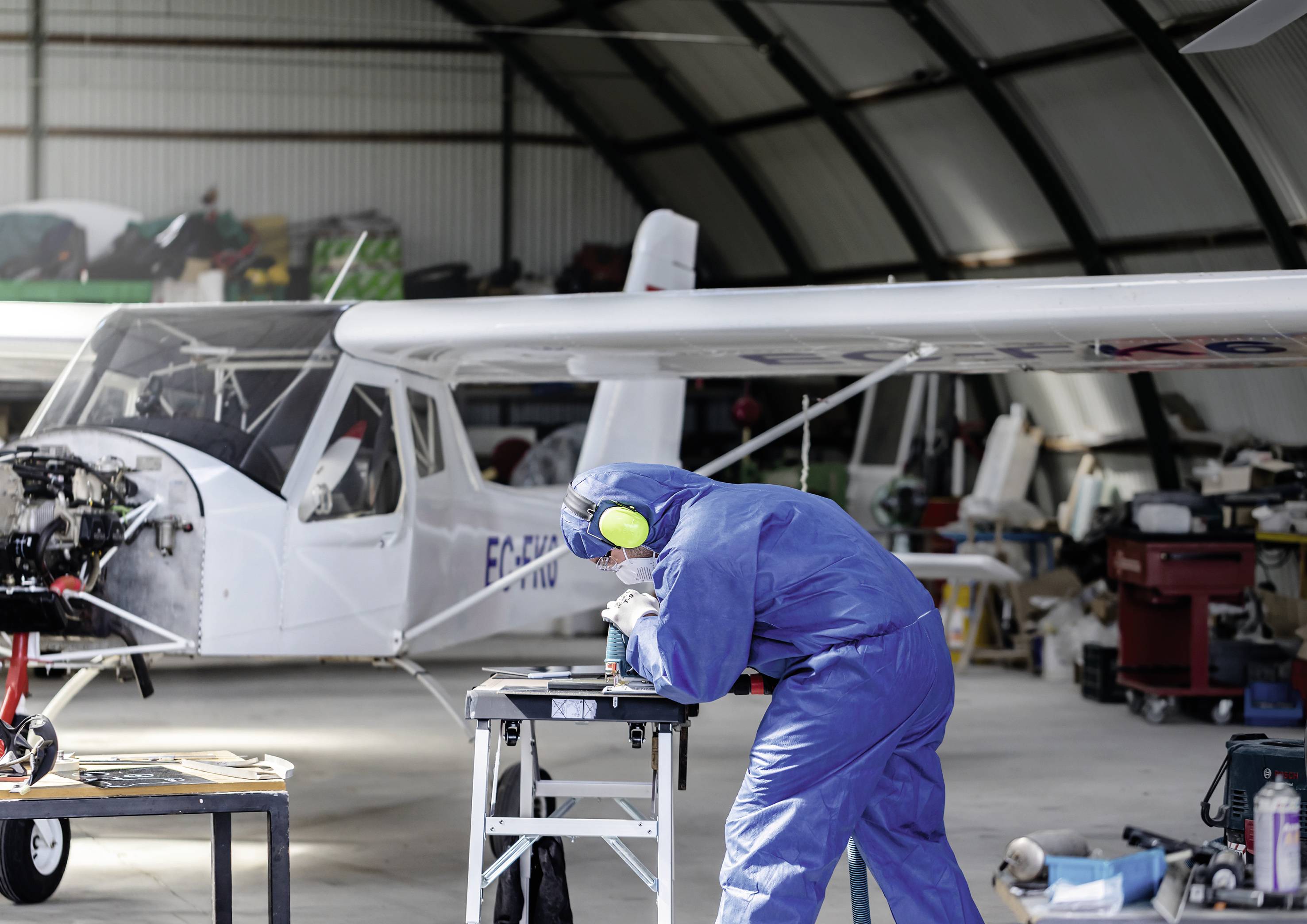 A technician in a blue protective suit is working at a workstation in a hangar. A small aircraft is visible in the background.