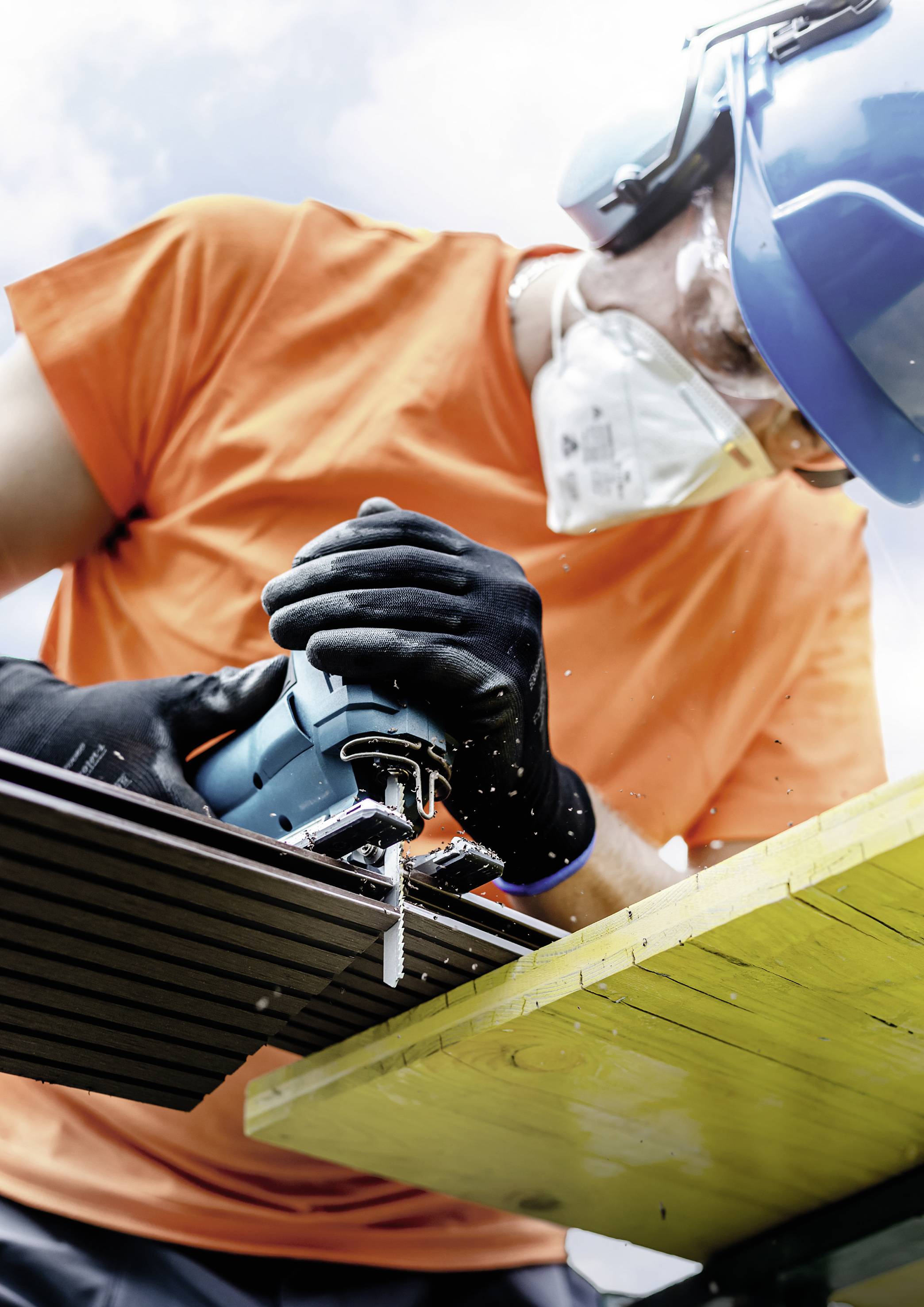 A person wearing protective clothing, a helmet, and a mask is sawing wood with an electric saw outdoors.