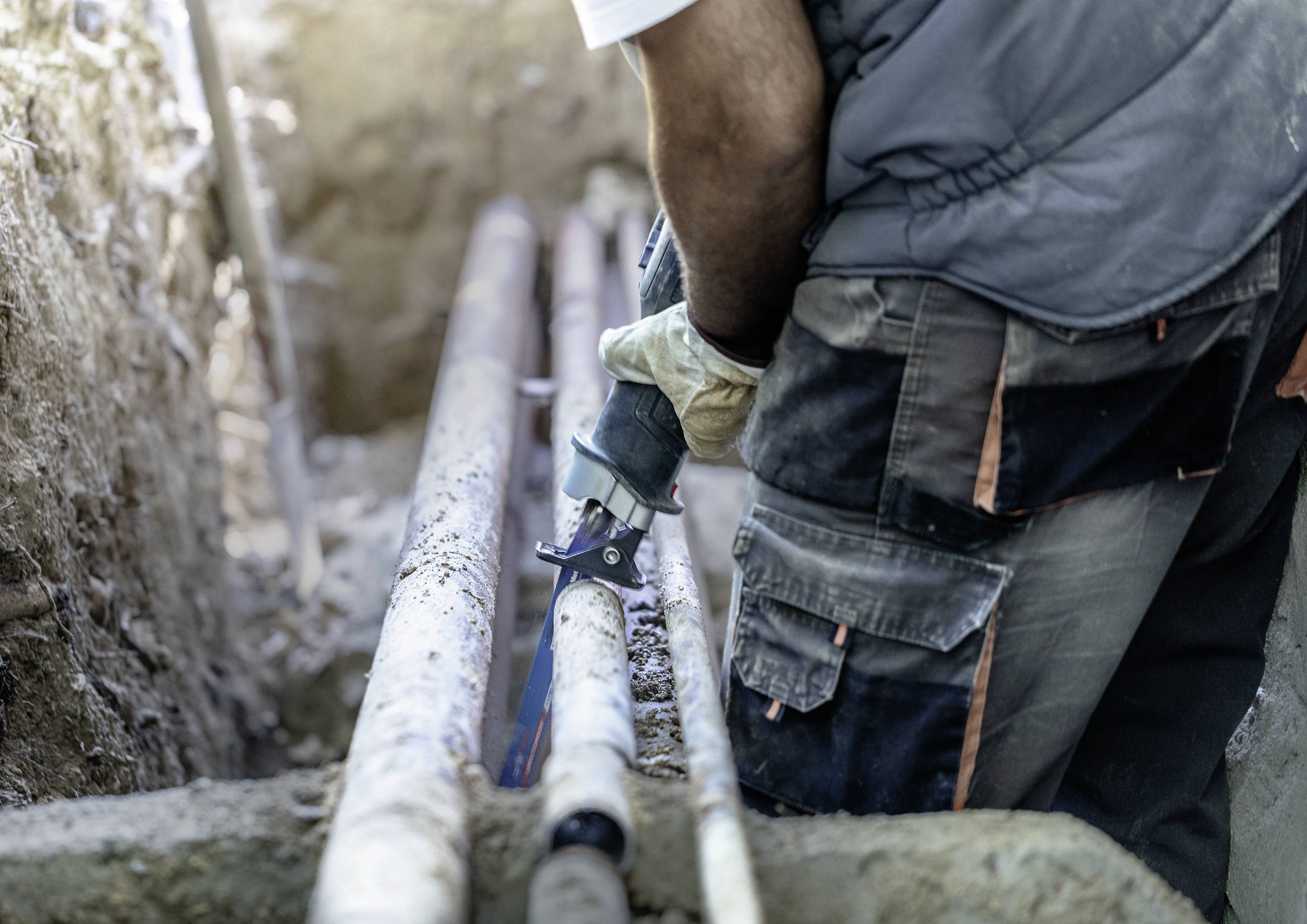 A worker is cutting a pipe with an electric saw. Surrounded by earth in an excavation pit. Gloves and protective clothing are being worn.