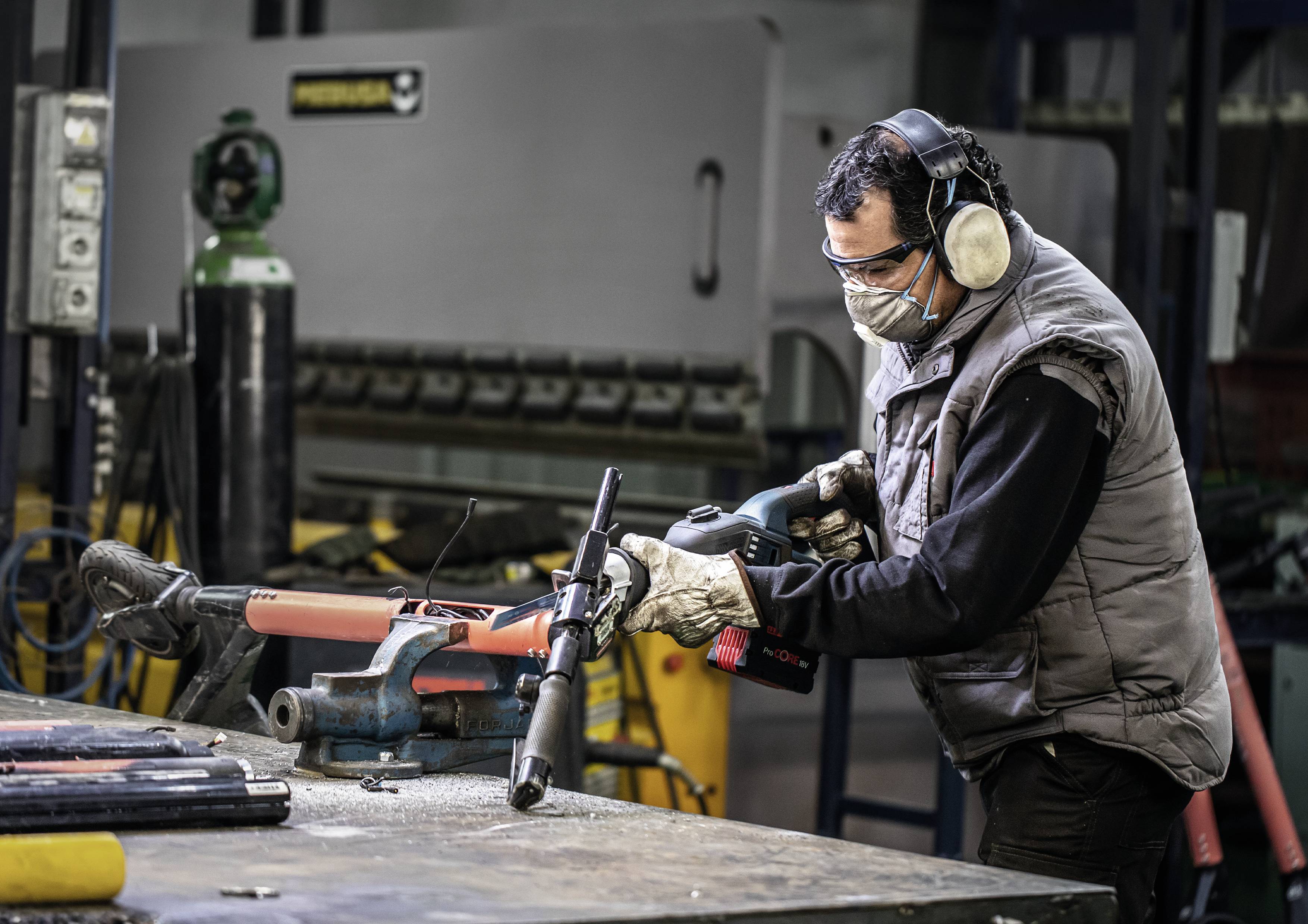 'Worker in workshop, wearing protective mask and ear protection, using an electric tool to work on metal pipe on workbench.'
