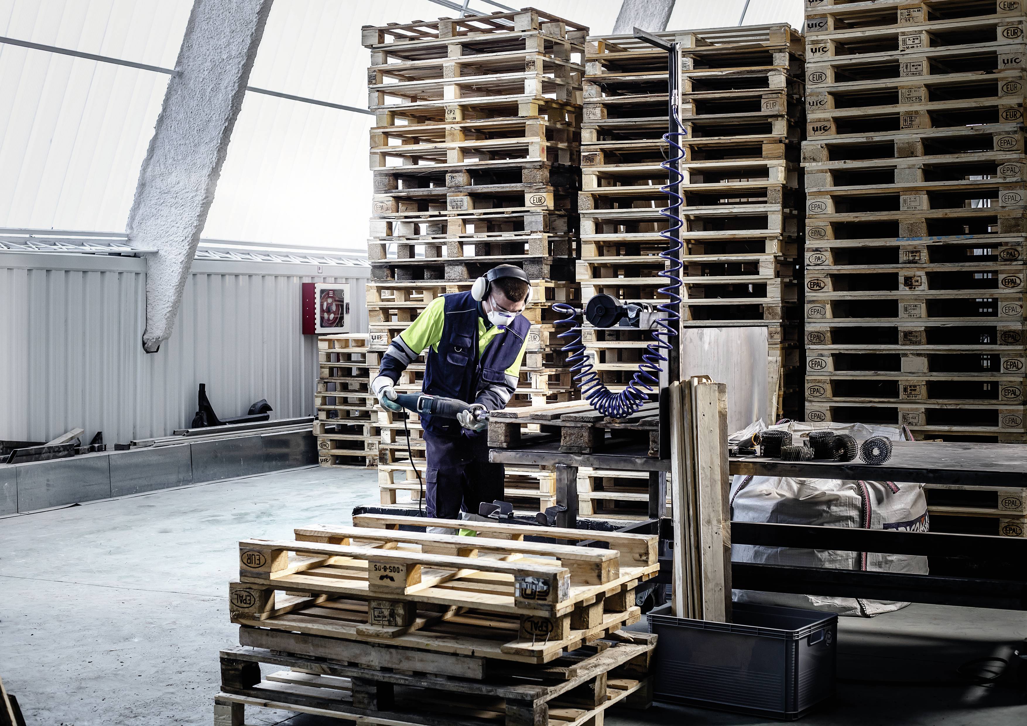 A person is working in a warehouse, sorting wooden pallets and using a device for handling the pallets.