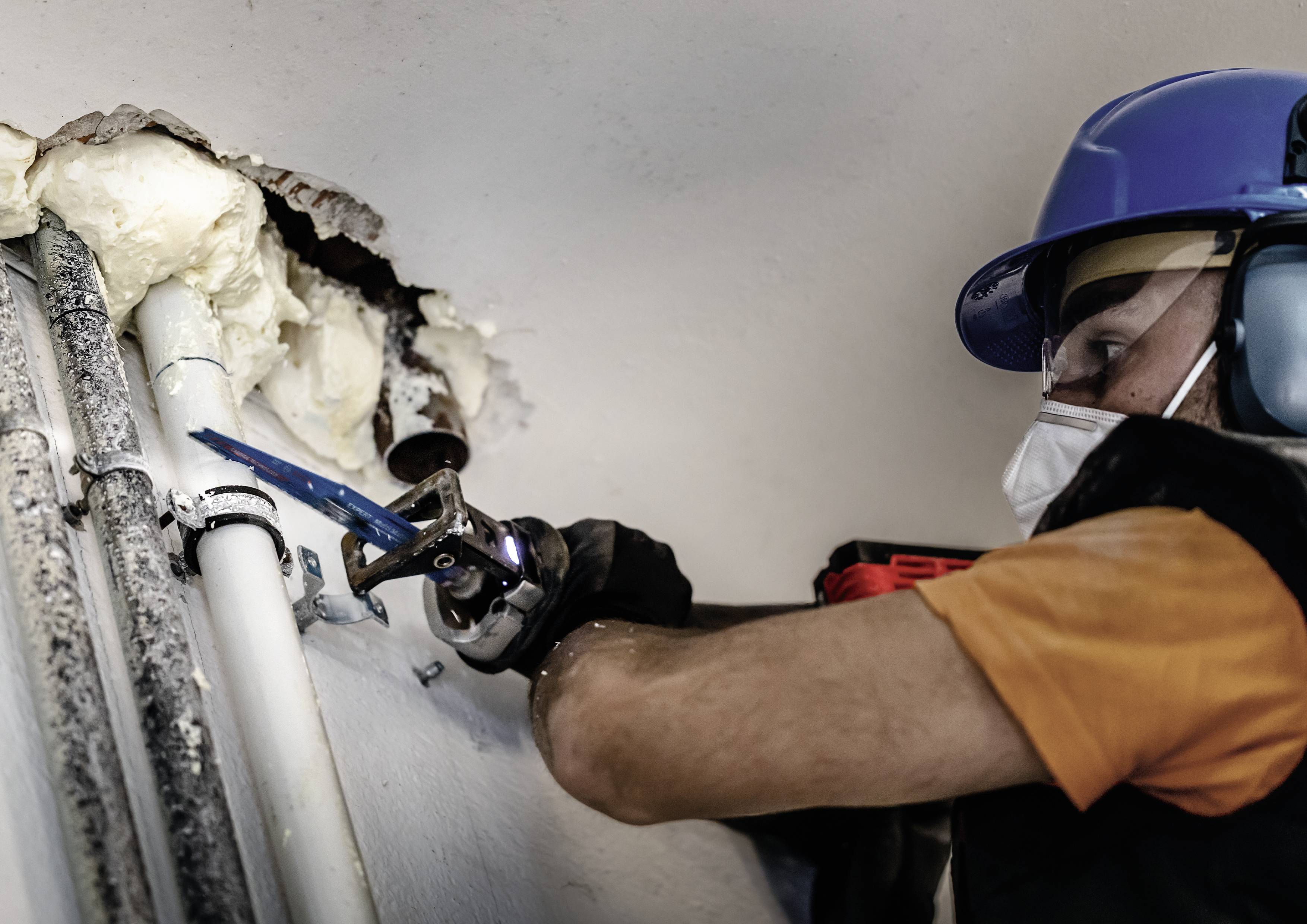 A plumber wearing a hard hat and mask is repairing pipes inside a wall. Foam insulation around the pipes is visible.