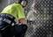A worker wearing safety clothing and headphones is using a drill on a metallic, diamond-shaped plate.