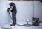 A worker in protective clothing is repairing a tile on the wall of an empty swimming pool. Next to him stands a vacuum cleaner with a long hose.