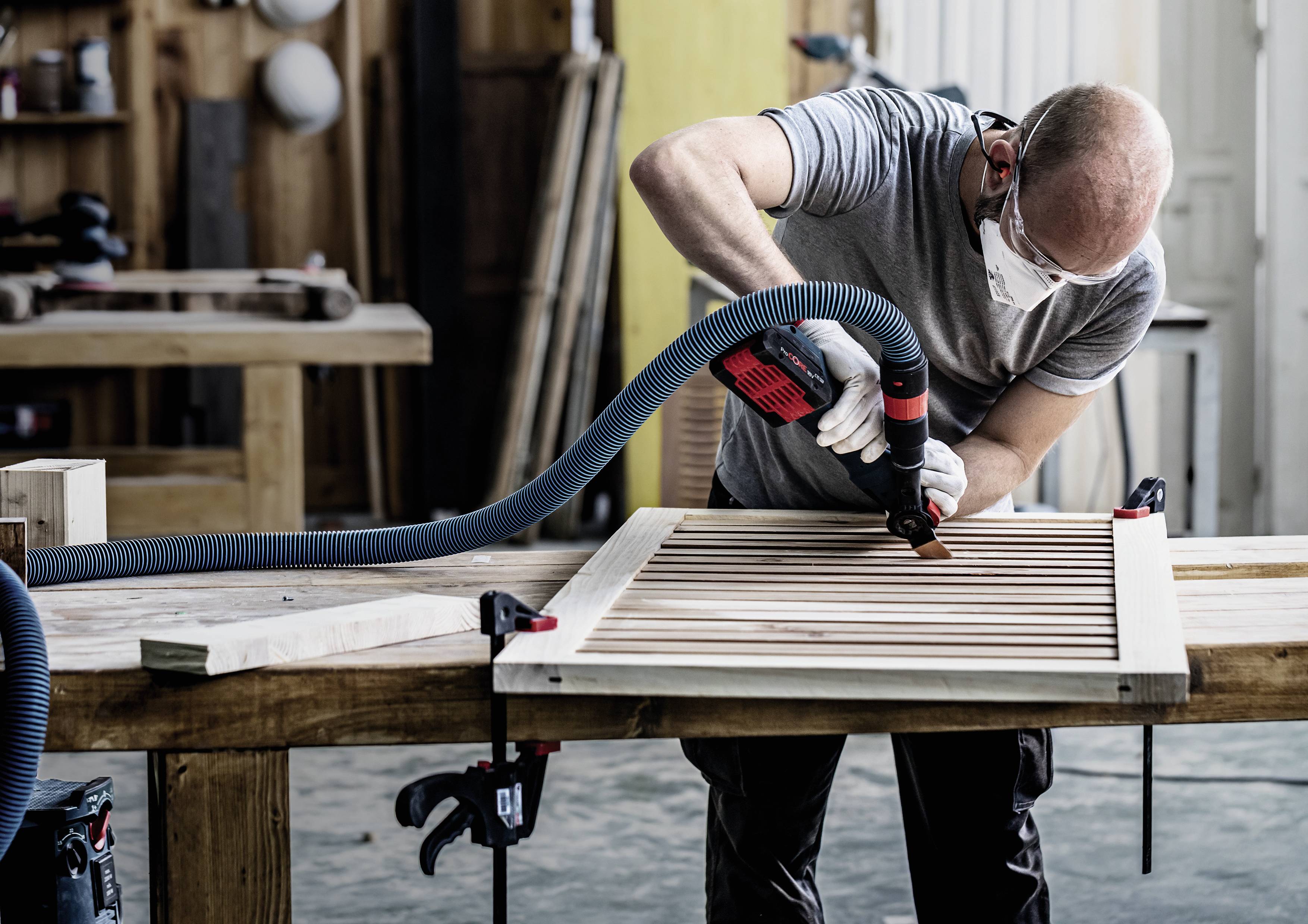 A man grinds a wooden board in a workshop, wearing a protective mask and using a grinding tool. Workshop tools and materials are visible in the background.