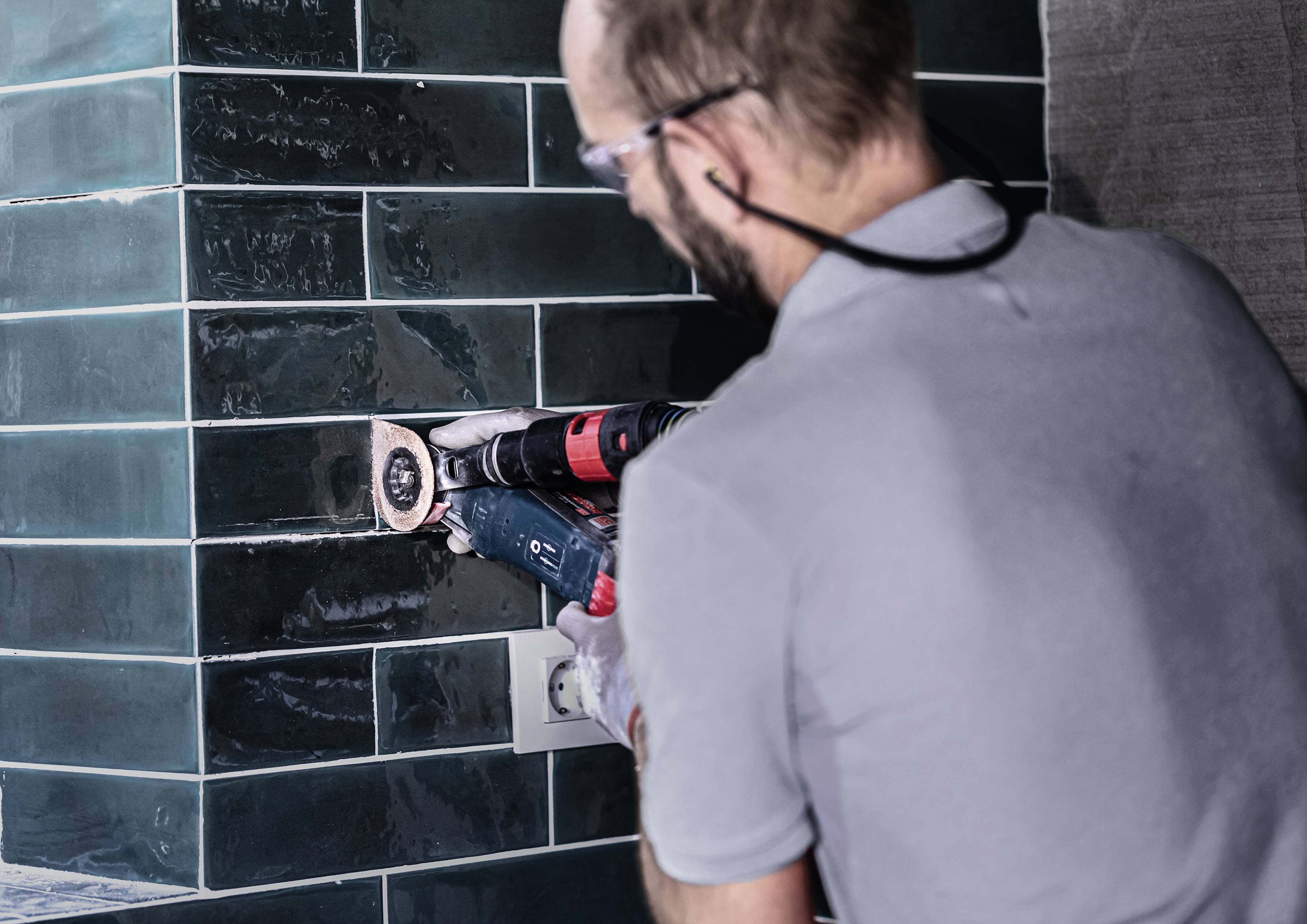 A person is wearing safety glasses and drilling into green tiles on a wall using an electric drill.