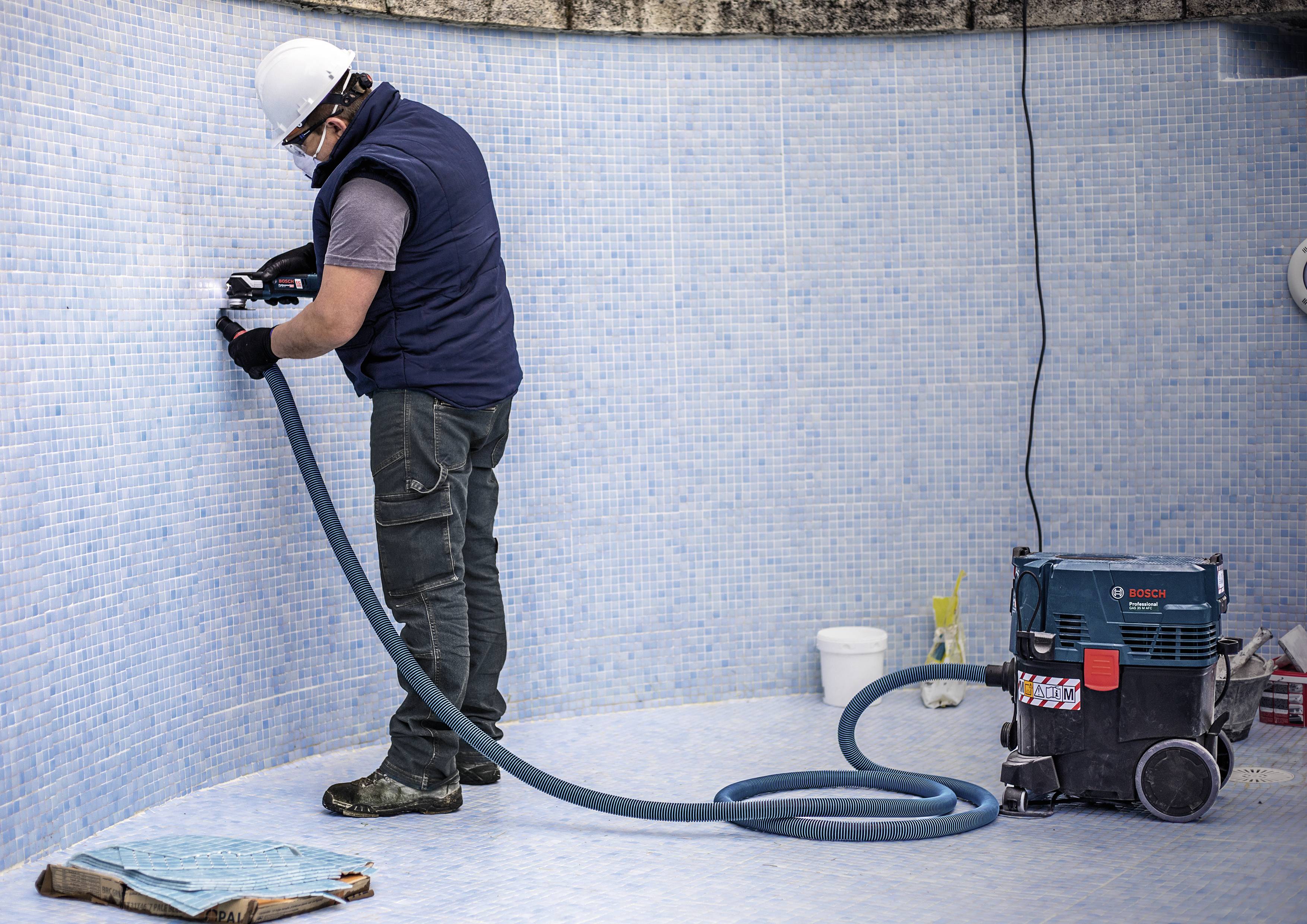 A worker wearing protective equipment is using an electric tool to clean or repair tiles in an empty swimming pool.