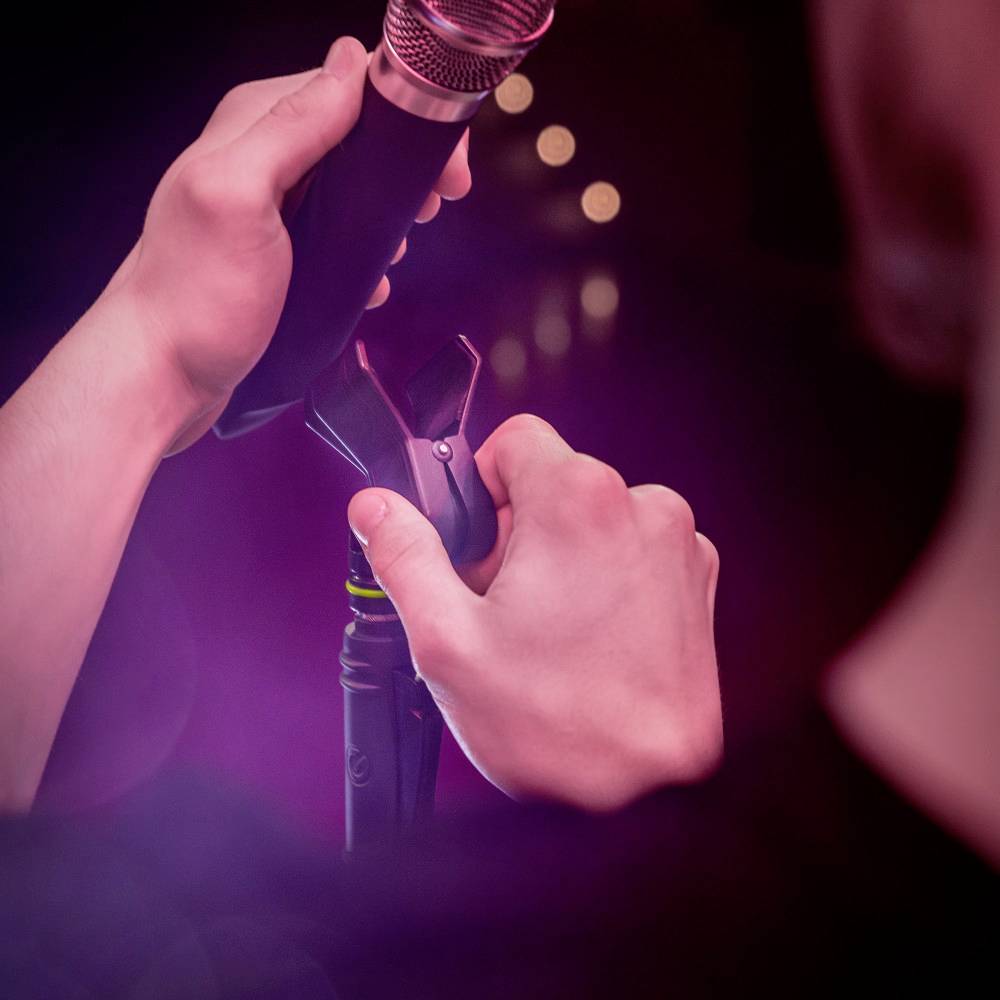 A person is holding a microphone that is mounted in a microphone stand. The background is out of focus and shows blurred lights.