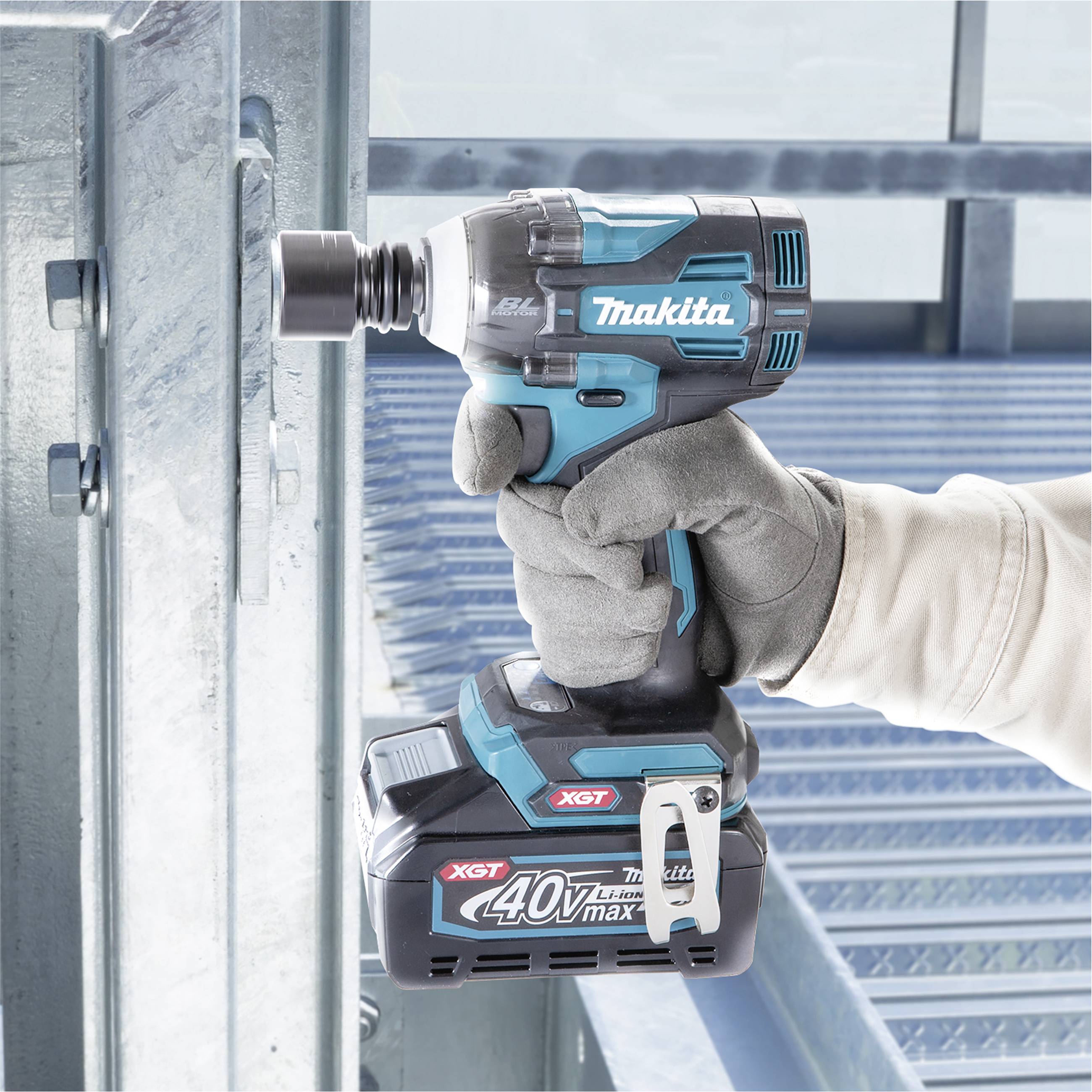 A person wearing a work glove is holding a blue cordless drill pointing towards a metal construction, with the 'Makita' logo visible.
