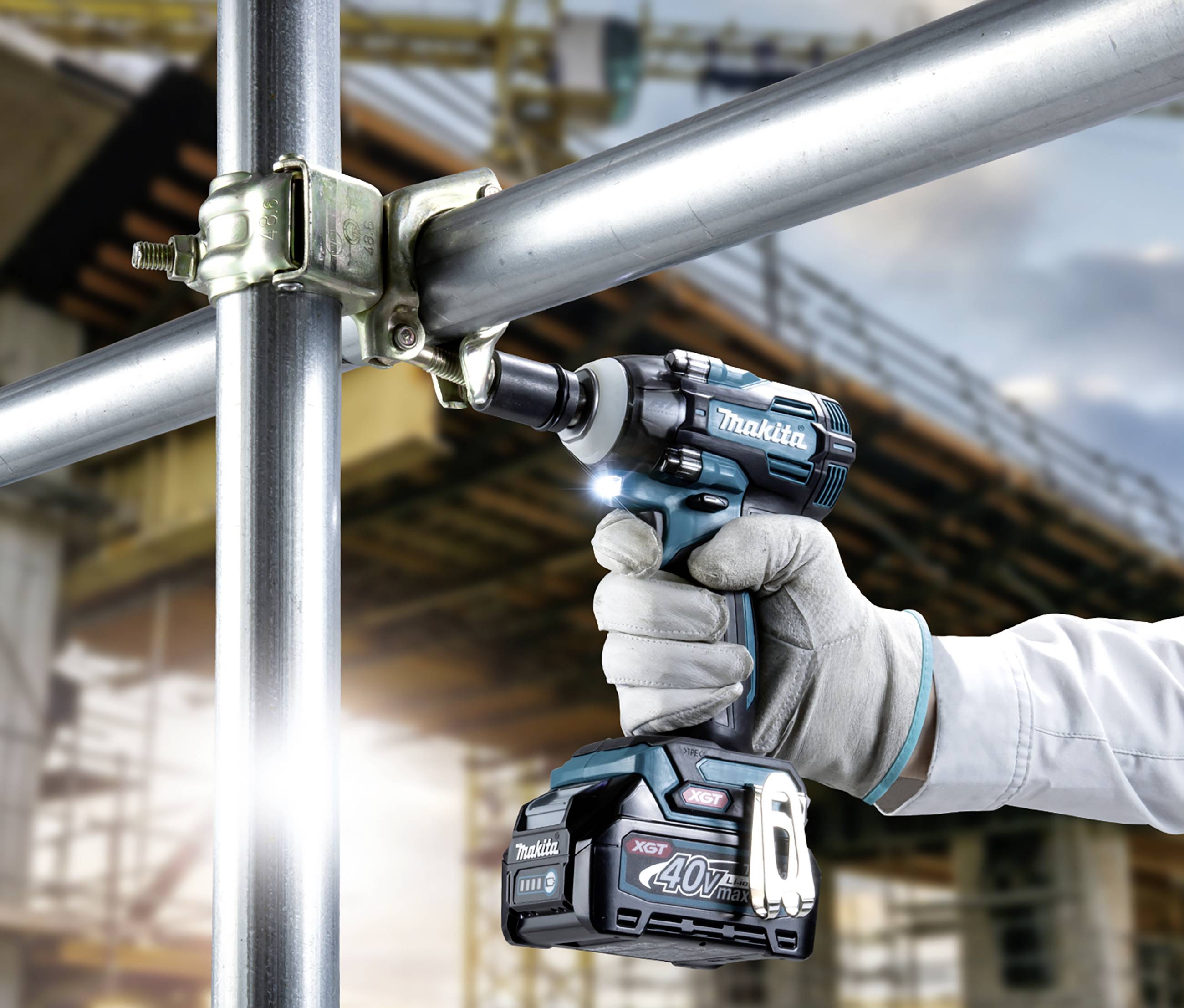 A person wearing work gloves is using a cordless drill to secure a scaffolding component, with a construction site in the background.
