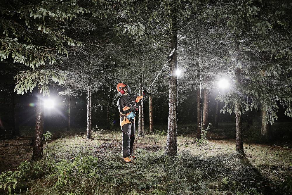 A person wearing protective clothing is cutting branches from a tree in an illuminated forest at night.