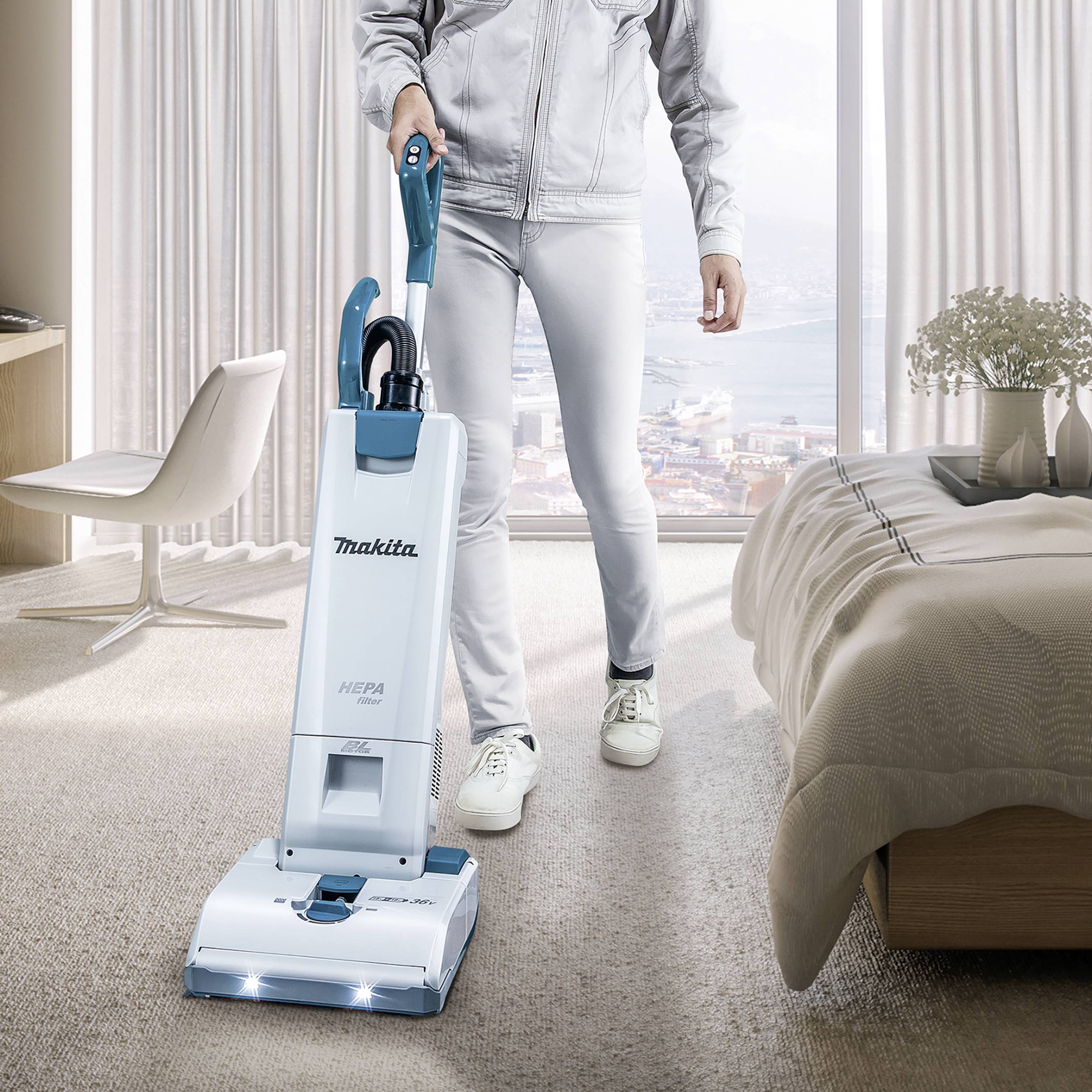 A person in a modern bedroom is using an upright vacuum cleaner. A window with a city view can be seen in the background.