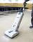 A person is using an upright vacuum cleaner to clean a carpeted floor in a large room with rows of chairs.