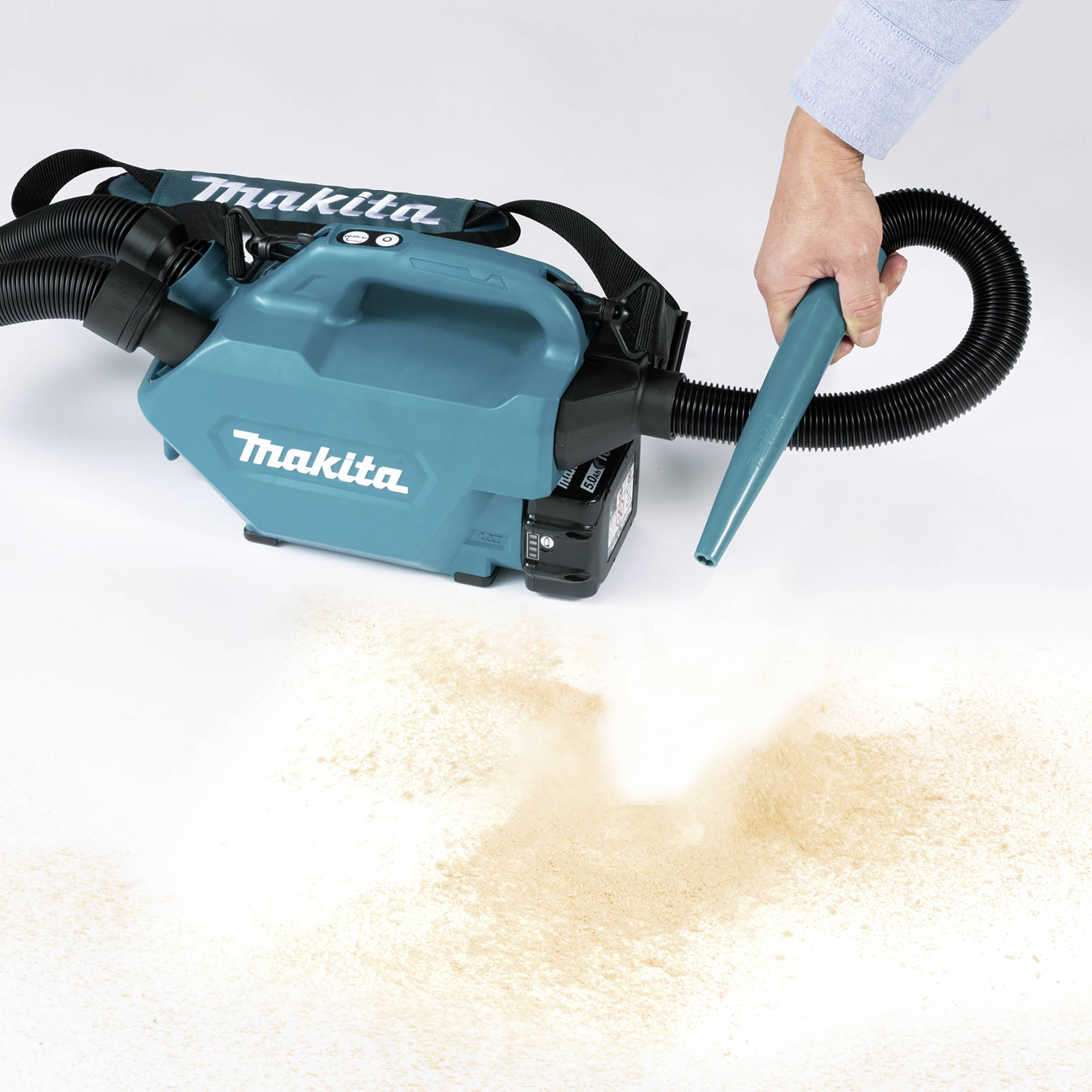 A Makita handheld vacuum cleaner is sucking up wood shavings. A person is guiding the hose over a surface with scattered wood chips.