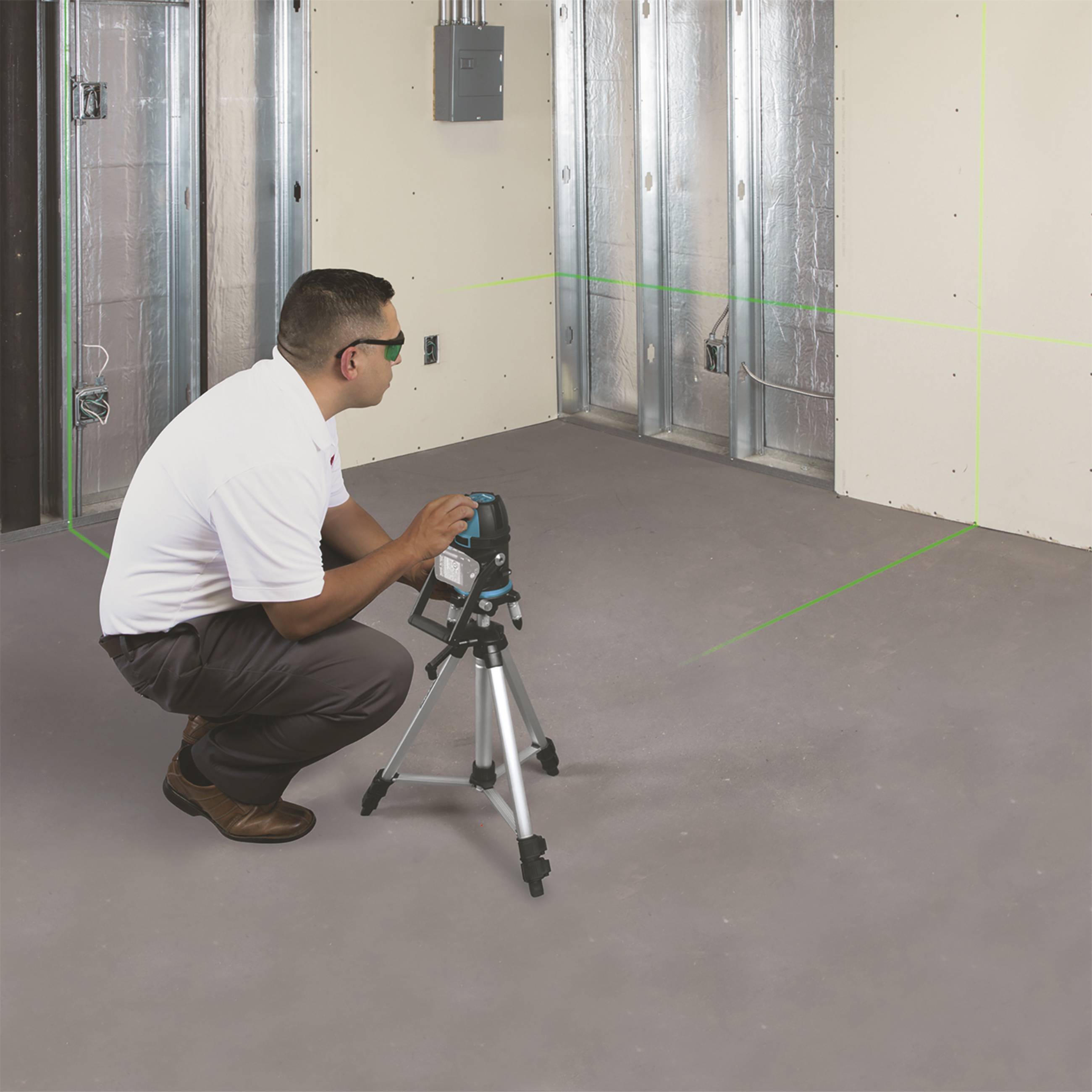 A man is aligning a laser levelling device on a tripod in a room under construction. Green lines mark the walls.