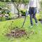 A person is standing in the garden using a leaf blower to gather leaves on the lawn. In the background, there are plants and a wooden fence.