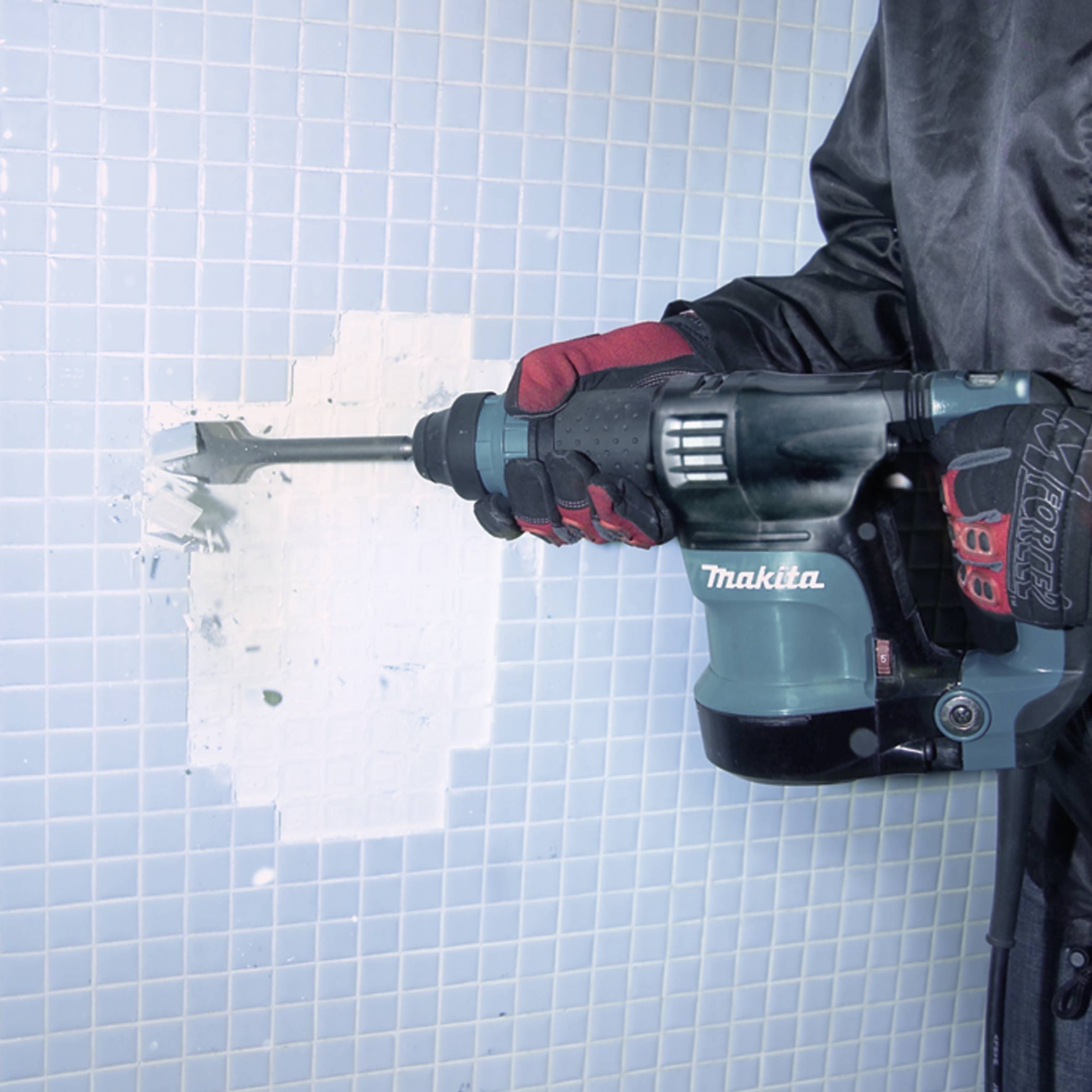 A person is using a hammer drill to remove white tiles from a wall. Small tile fragments are flying off.