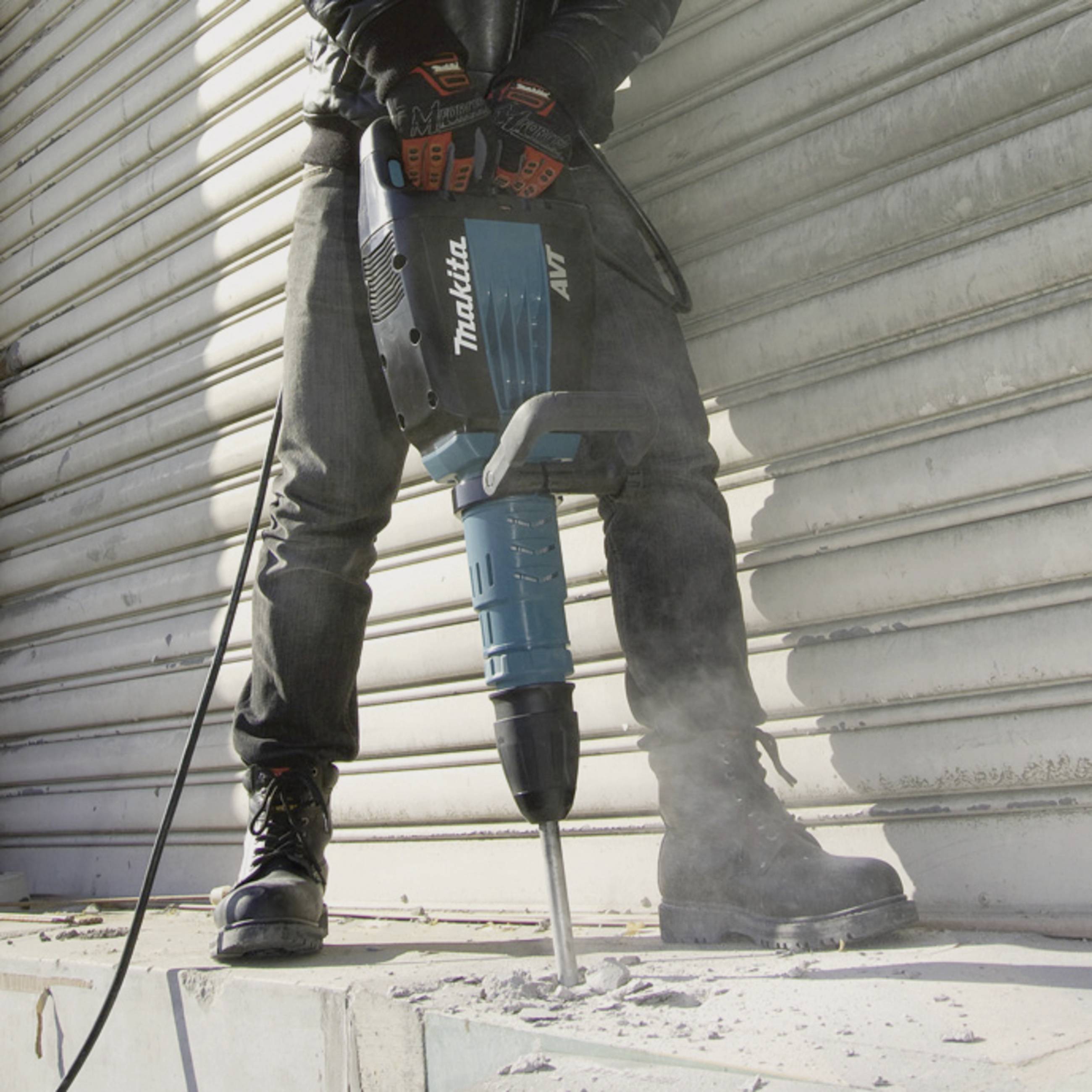 A person is using a blue impact drill to break concrete. The drill bears the 'Makita' logo.