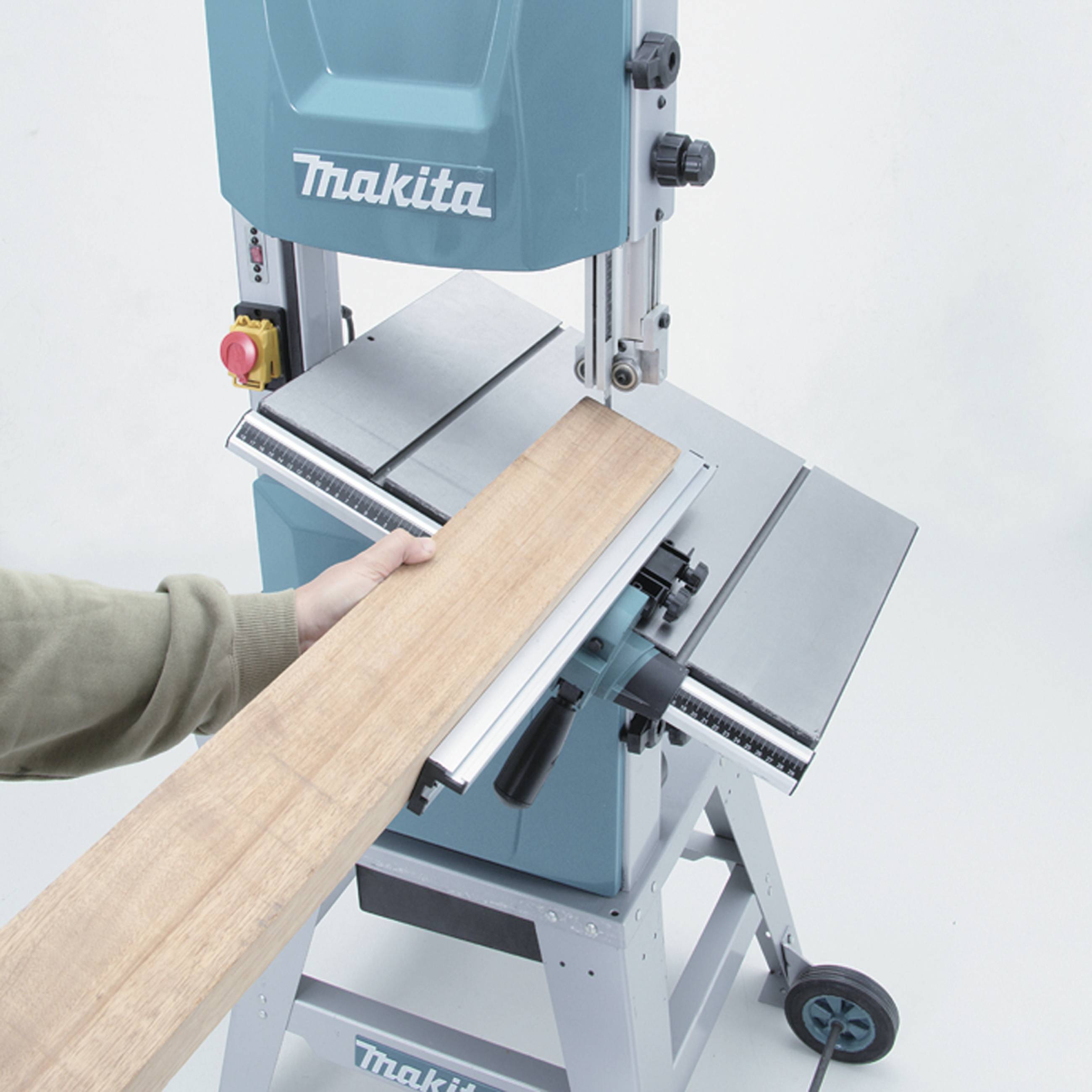 A person is cutting a wooden board on a blue bandsaw. The machine has an emergency stop button and metal guide rails.