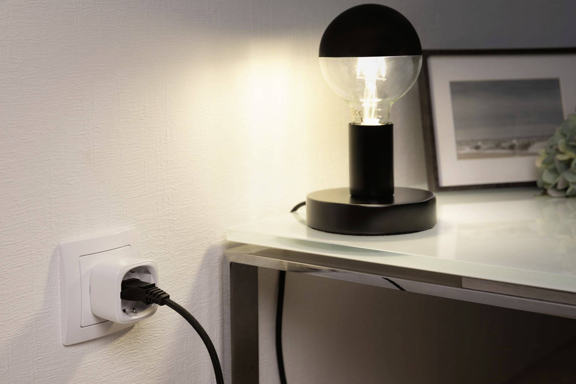 A table lamp with a clear light bulb design illuminates a table. In the background, a socket with a plug inserted is visible.