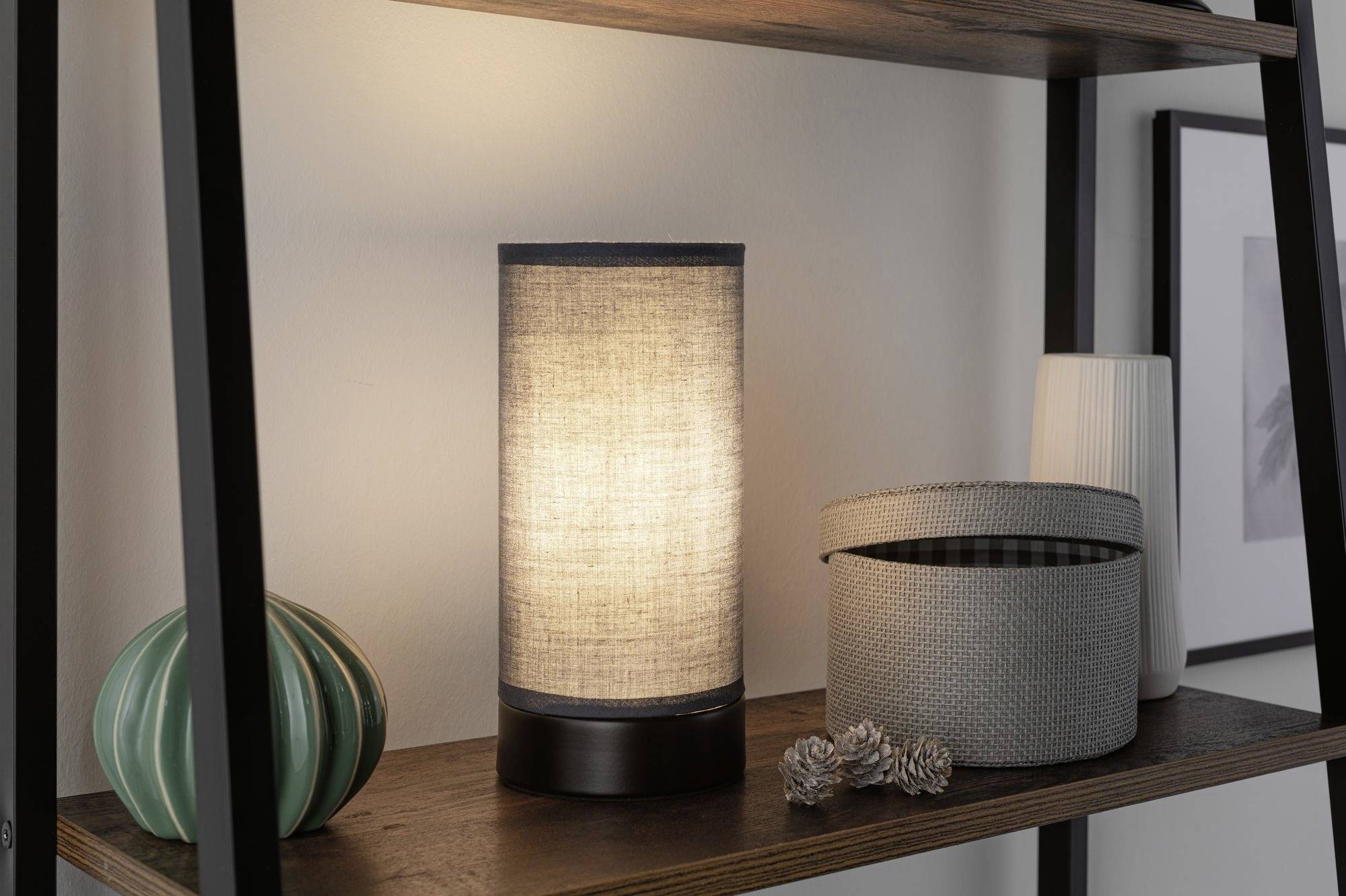 On a shelf sits a glowing table lamp with a fabric shade, next to decorative items like a green ceramic cactus and an open fabric storage box.