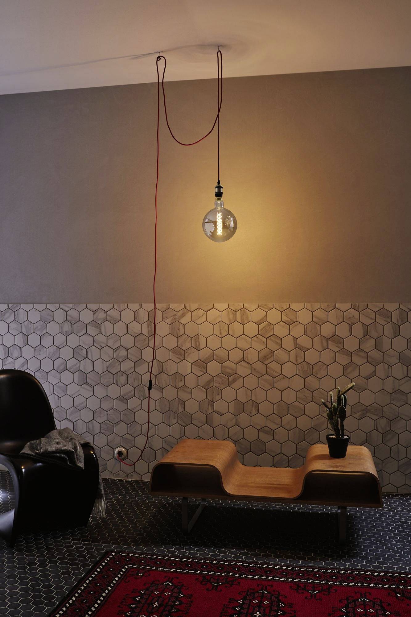 A stylishly decorated room with a hanging light bulb, a wooden daybed, a small plant pot, and geometric wall patterns.