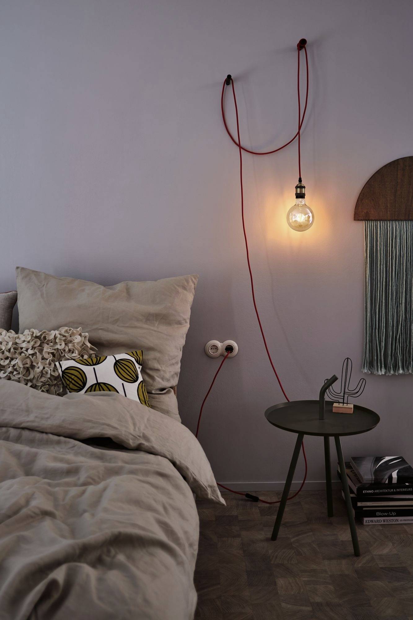 Cosy bedroom with a bed adorned with cushions. A hanging light bulb on the wall to the left. A side table with a cactus decoration underneath.