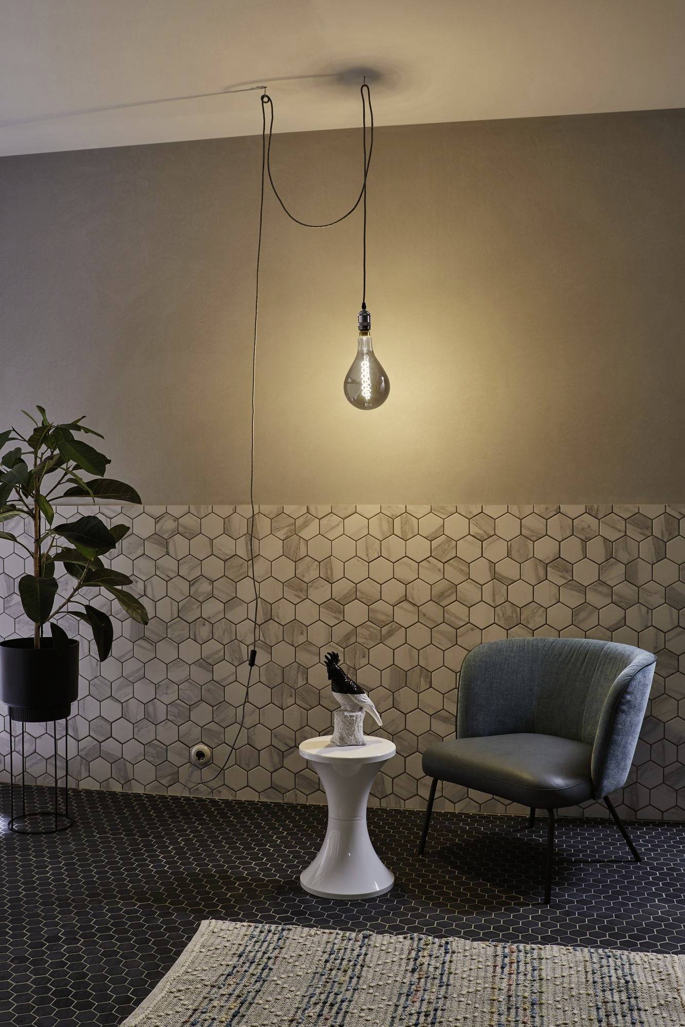 A stylish room with a hanging light fixture, an armchair, a small table and a potted plant in front of a decorative wall.