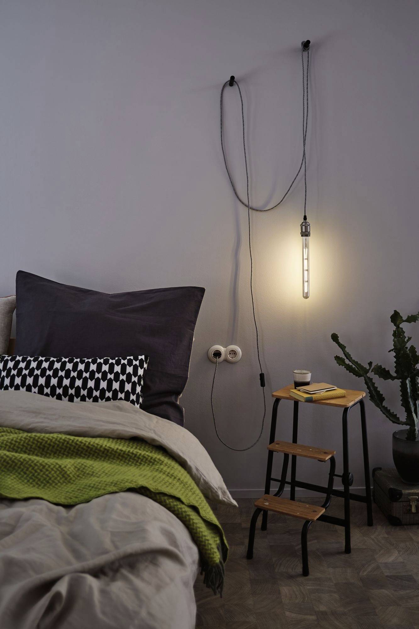 A bedroom with a hanging light bulb on the wall, a cosy bed with a green blanket, and a small stool.