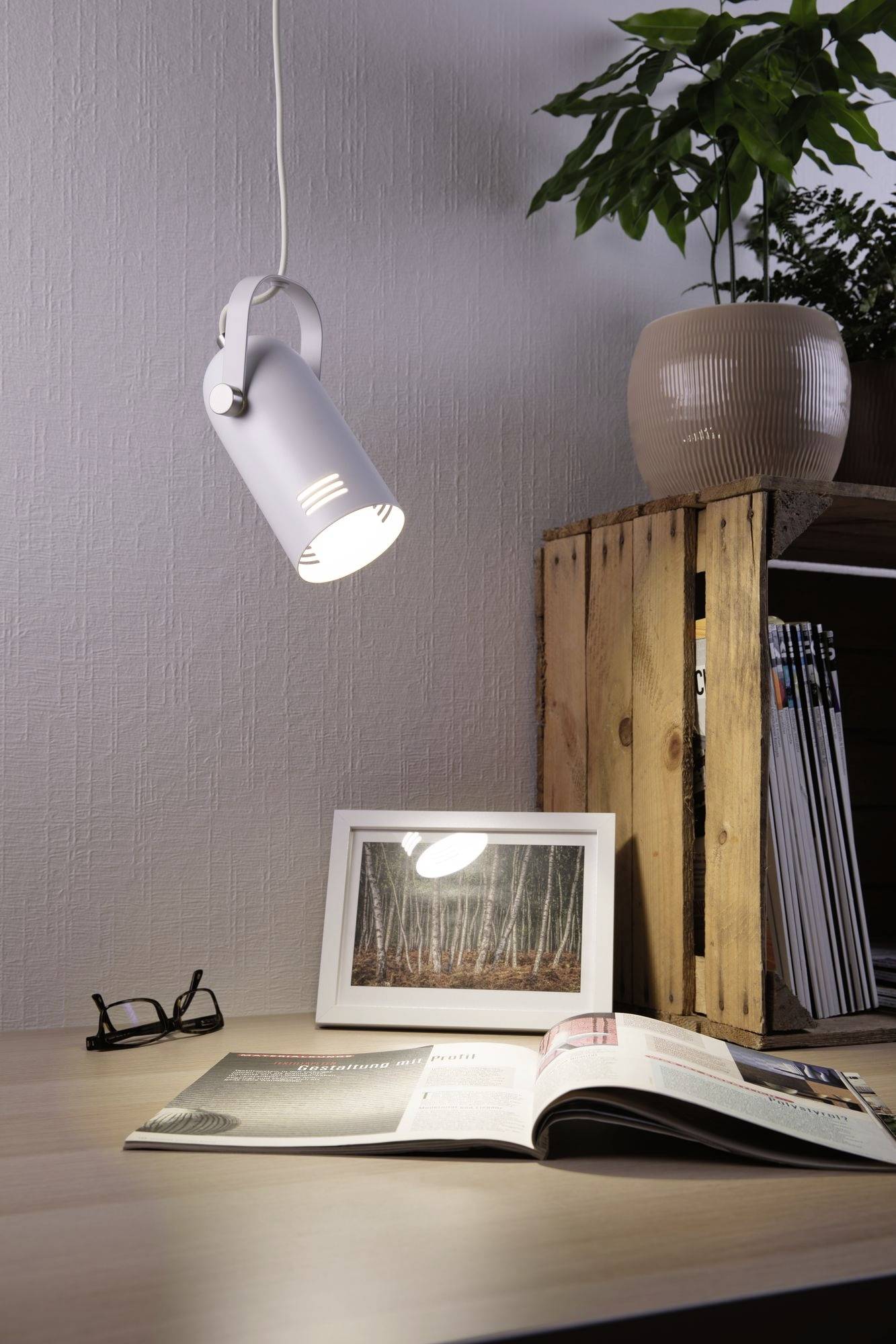 A white lamp illuminates a picture of trees on a desk. Next to it are a pair of glasses, an open magazine and a plant pot.
