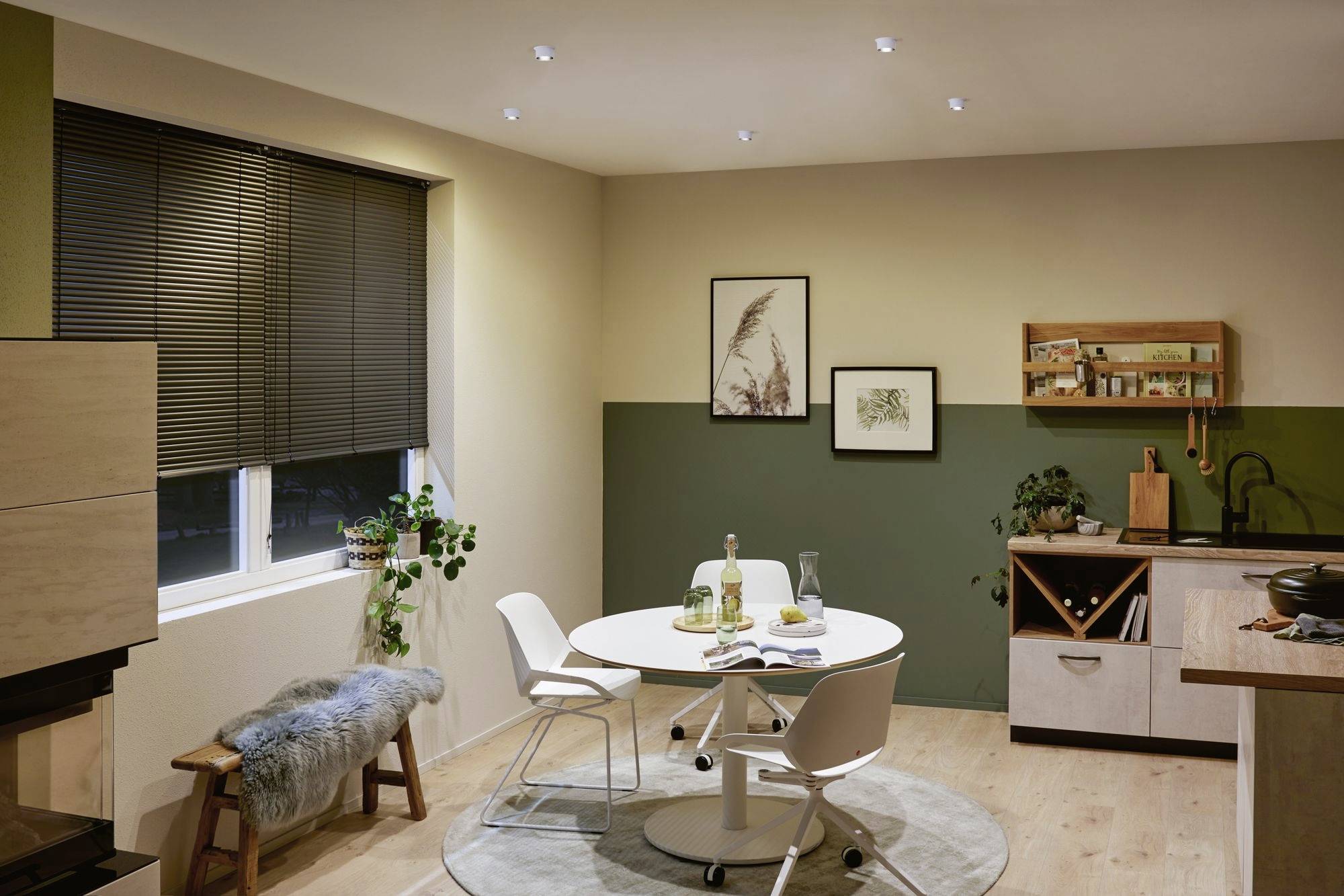 A modern dining room with a round white table and four chairs. In the background, plants and framed pictures are displayed on the wall.