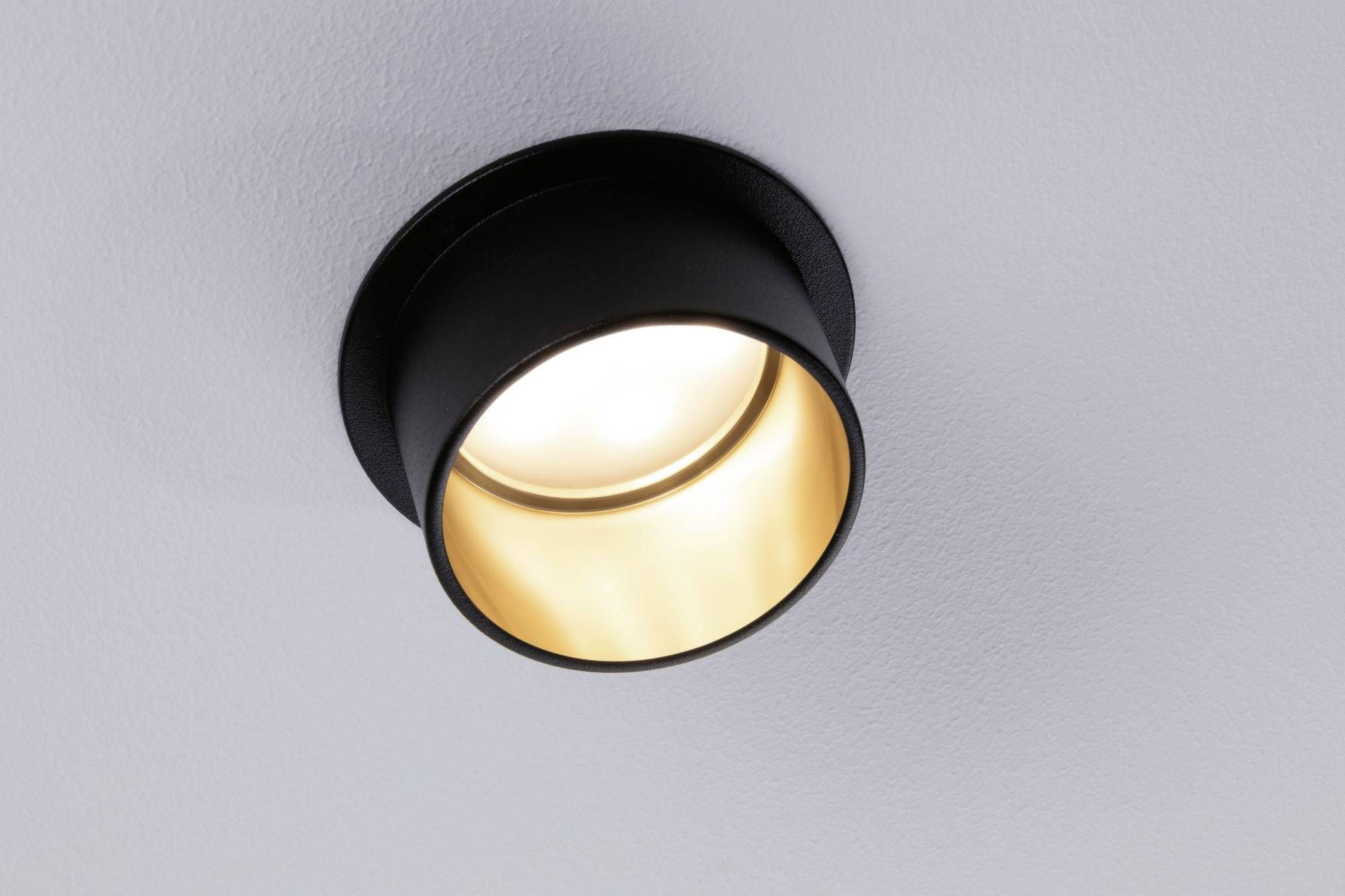 Black ceiling light with a gold-coloured interior, casting light downwards. Mounted on a white ceiling.