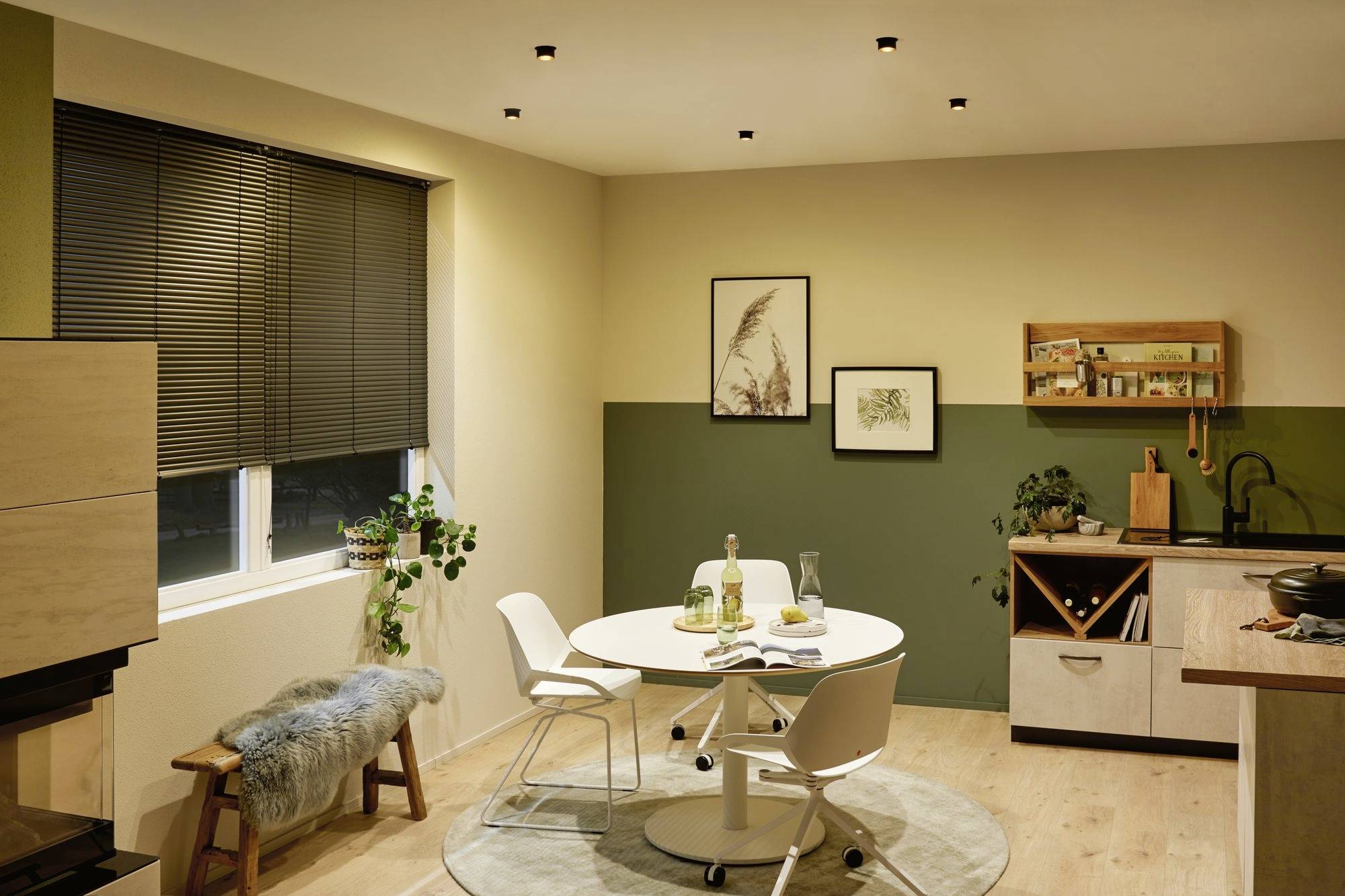 Modern kitchen with a round table and four chairs. Dark green and cream walls, plants, shelving, and artwork on the walls.